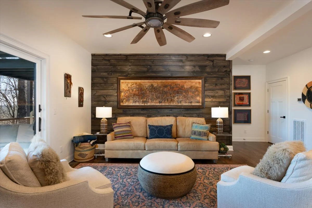 Living room with a beige sofa, cream armchairs, a round ottoman, and decorative pillows. Dark wood accent wall with a framed painting, flanked by two lamps. Ceiling fan with wooden blades, sliding glass door to the balcony, and framed artwork on the walls.
