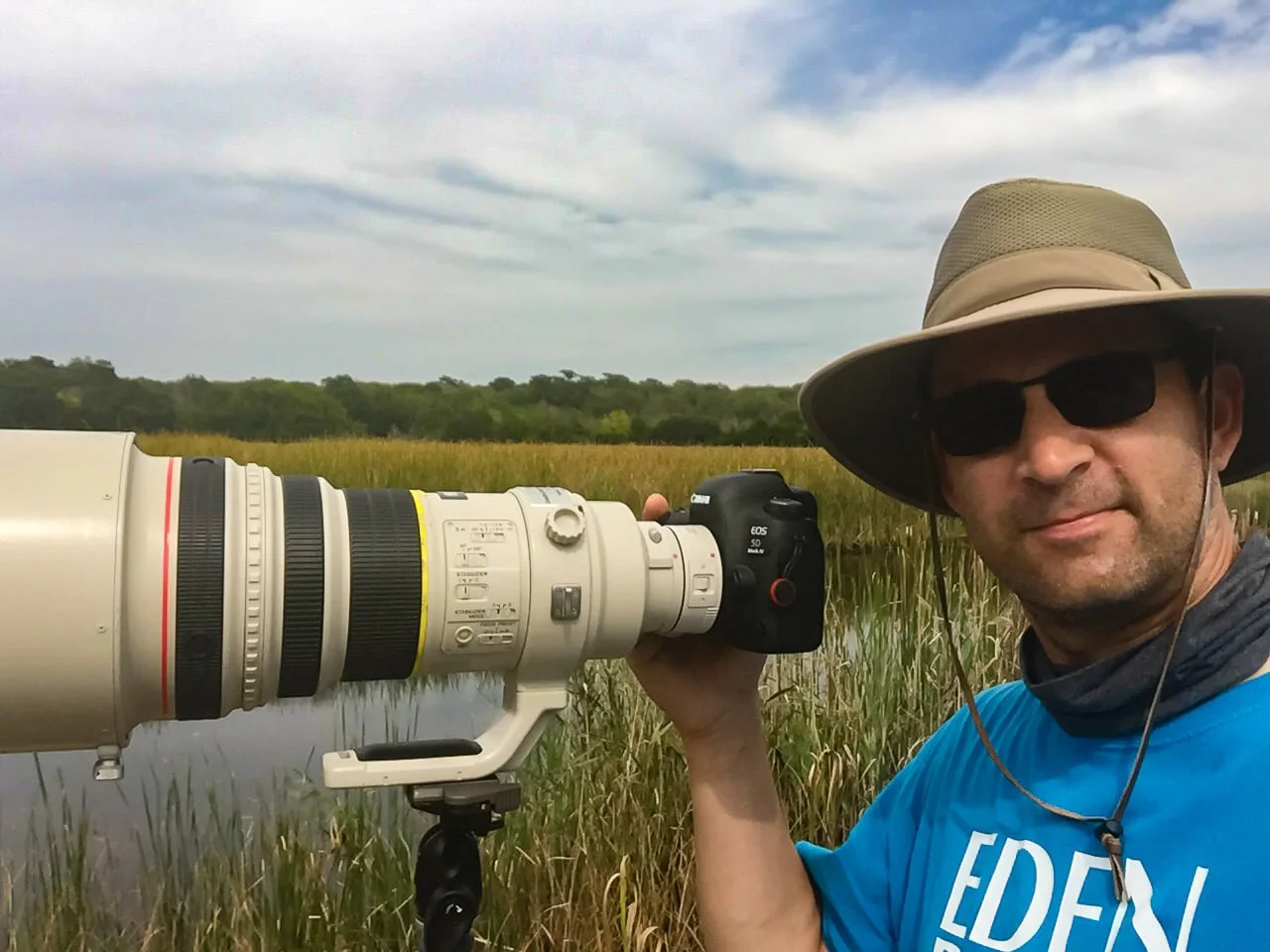 A man wearing a wide-brimmed hat, dark sunglasses, and a blue shirt holding a professional camera with a long telephoto lens outdoors near wetland vegetation, with a cloudy sky in the background.