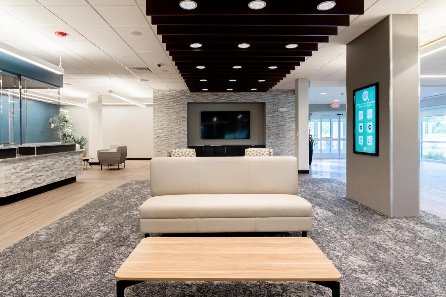 Modern lobby area with a beige couch, a wooden coffee table, a TV mounted on a grey brick wall, and a digital display on the right wall. There are chairs and a reception desk on the left, with large windows and glass doors in the background.