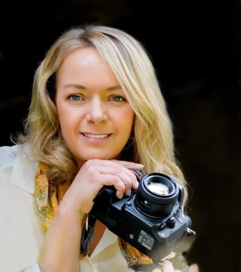 A woman with blonde hair holding a camera, smiling at the camera against a black background.