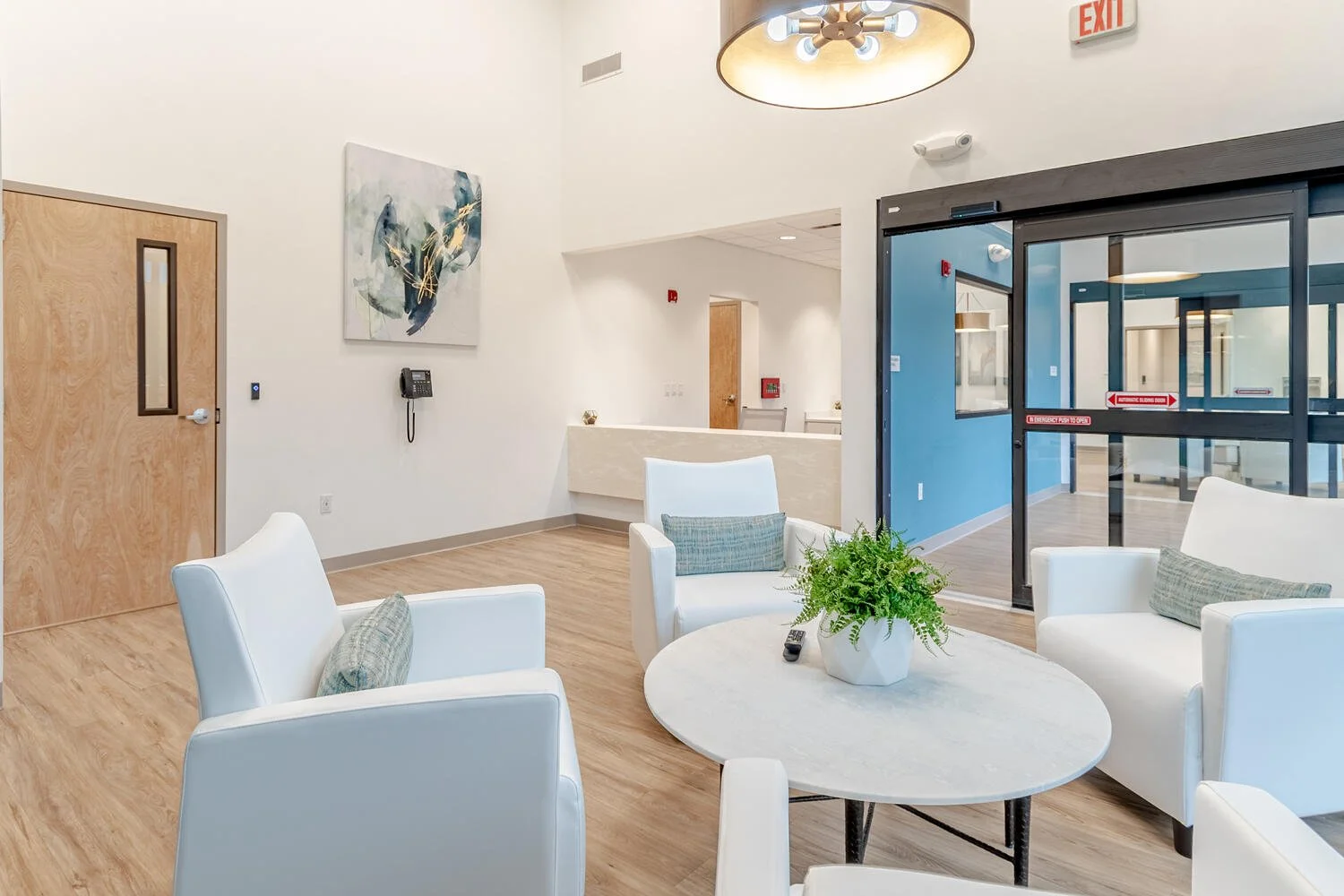A modern waiting room with four white armchairs around a round table with a potted green plant and remote control, against a white wall with decorative art, a mounted telephone, and a door leading to another room.