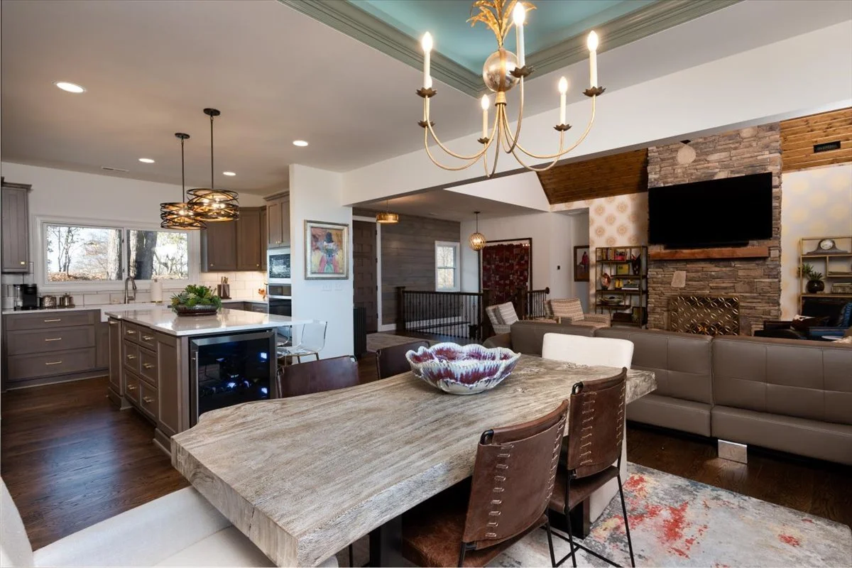 Open-concept living and kitchen area with wooden dining table, modern chandelier, stone fireplace, and contemporary furniture.