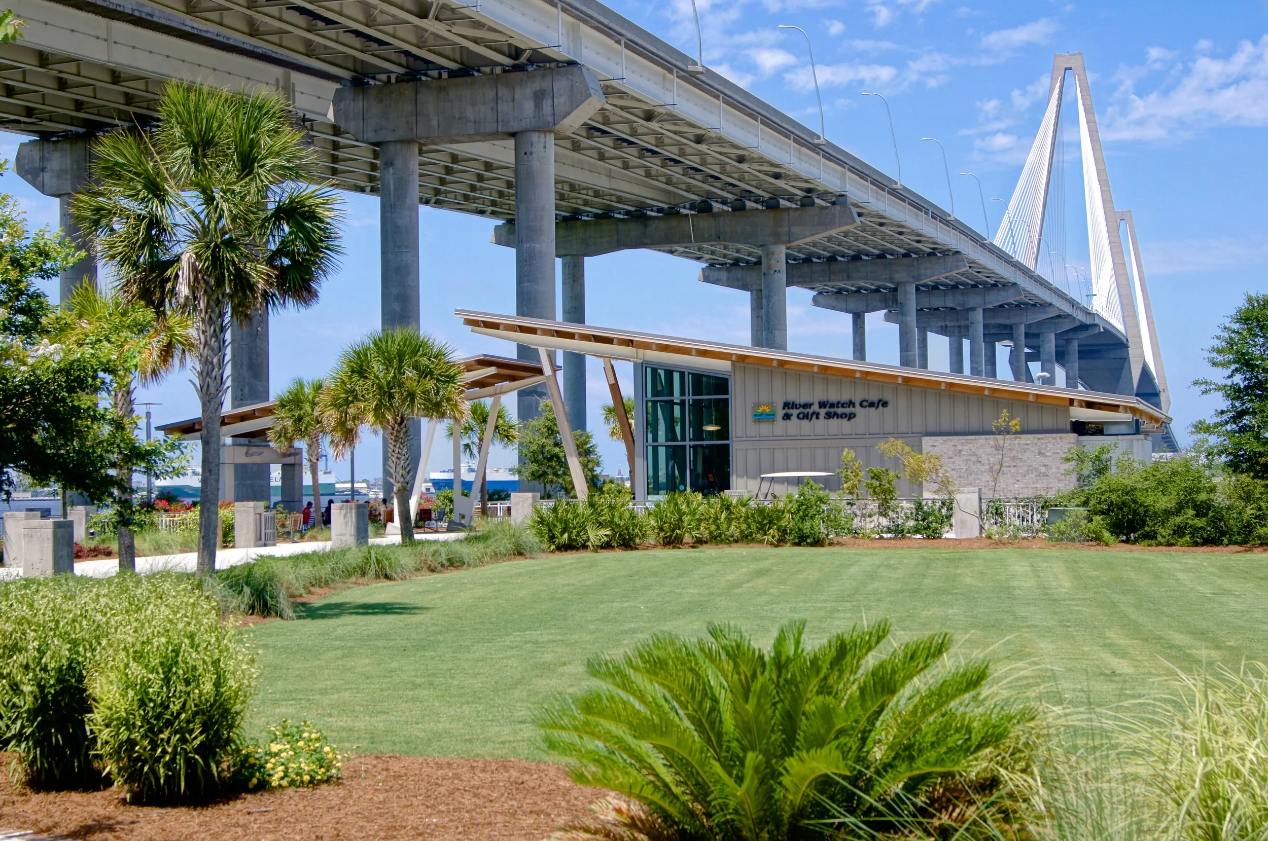 View of a park with green grass, palm trees, and shrubs in front of a bridge with concrete pillars and a modern design, under a blue sky with some clouds. A building with a sign reading 'River Watch Cafe & Gift Shop' is nearby.
