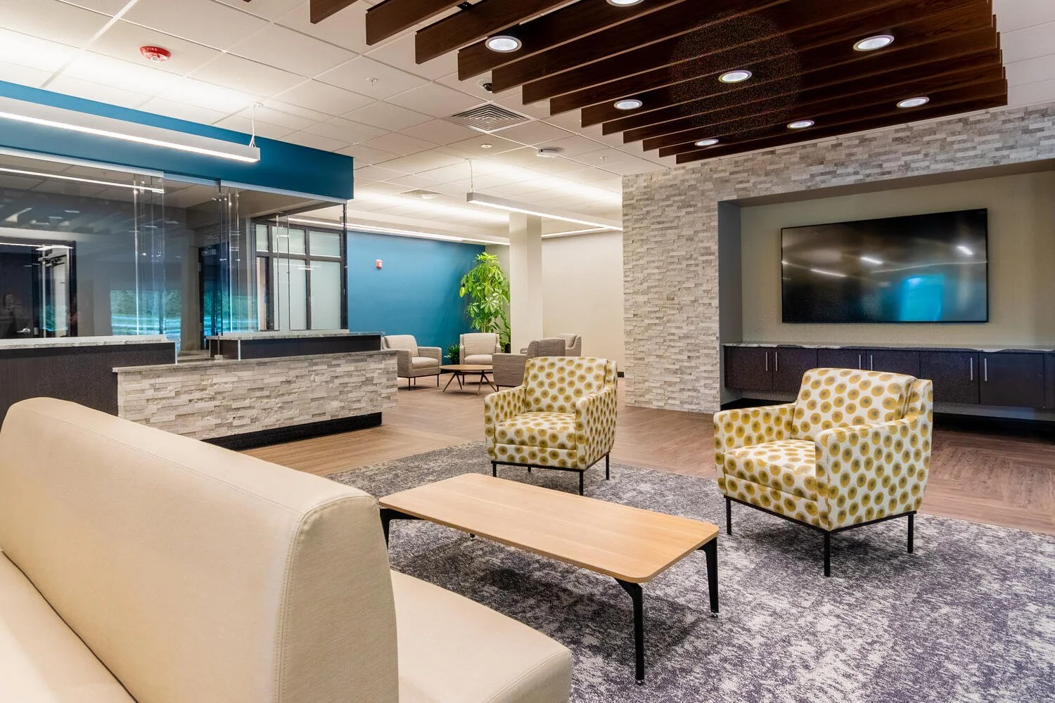 Modern lobby with beige sofa, yellow patterned armchairs, wooden coffee table, gray area rug, stone accent wall, mounted flat screen TV, and a seating area with additional chairs and a large plant.