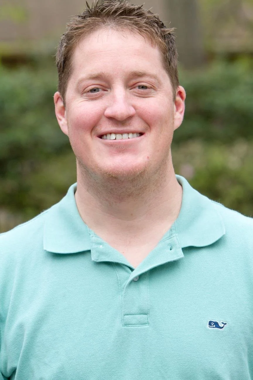 A smiling man with short, light brown hair wearing a light blue polo shirt outdoors with blurred green trees in the background.