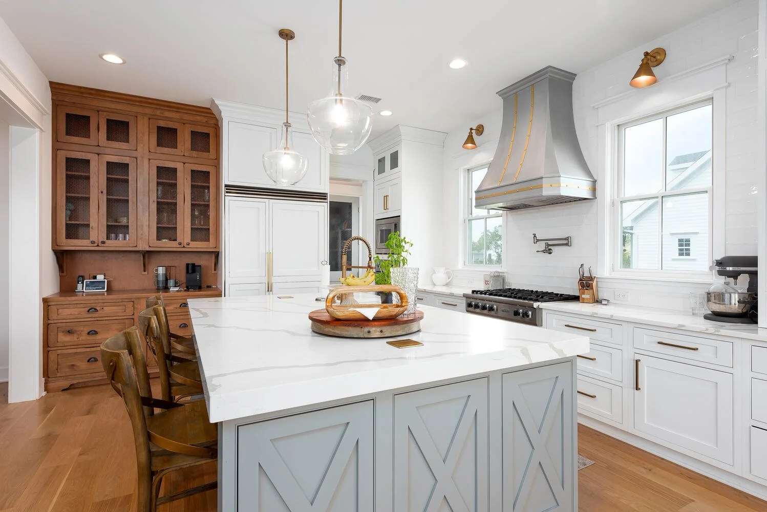 Modern kitchen with white cabinetry, a large island with a marble countertop, wooden chairs, stainless steel range hood, and natural light from two large windows.
