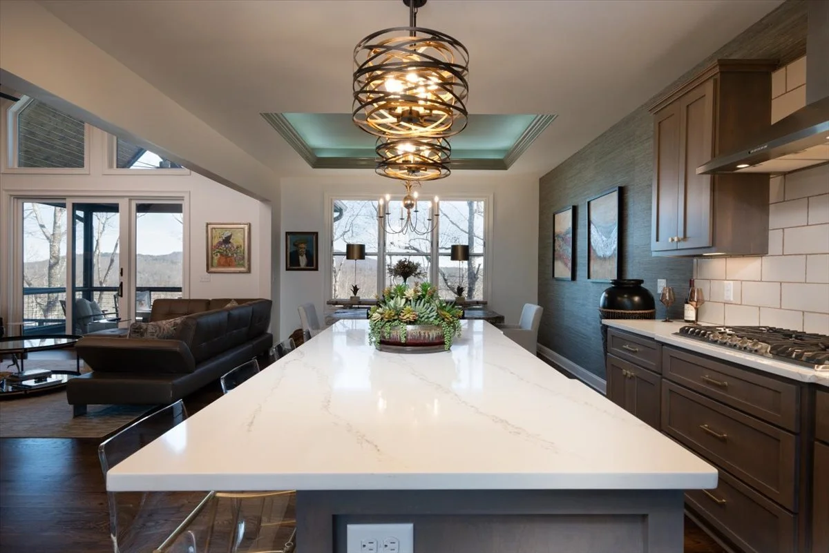 Modern kitchen with a large white island, pendant and chandelier lighting, wooden cabinetry, and a living room with a black sofa in the background.
