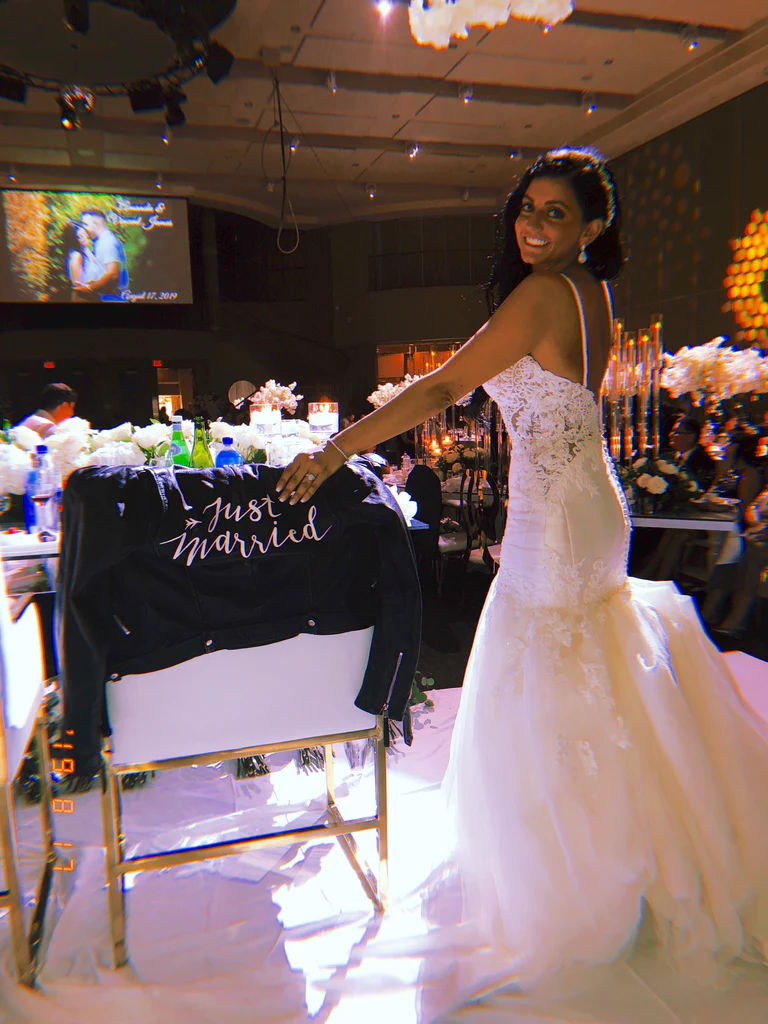 A bride smiles for the camera with her "just married" leather jacket hanging on her chair.