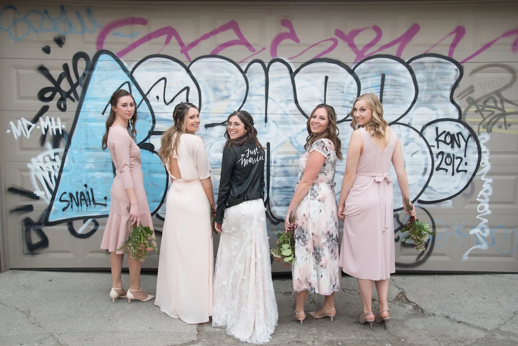A bride and her bridesmaids pose for a photo in front of some graffiti.