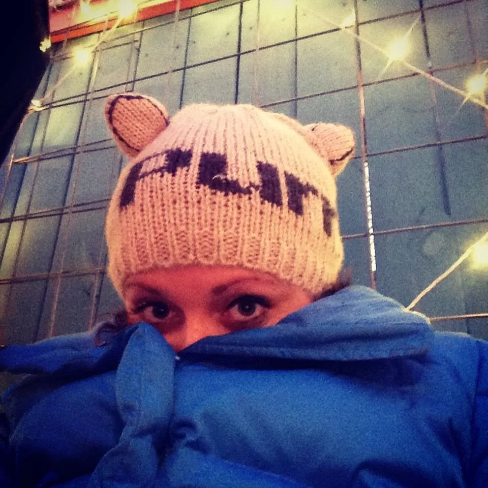 A person wearing a knitted hat with cat face and ears, and a blue jacket, partially hiding their face, with a metal grid background and string lights.