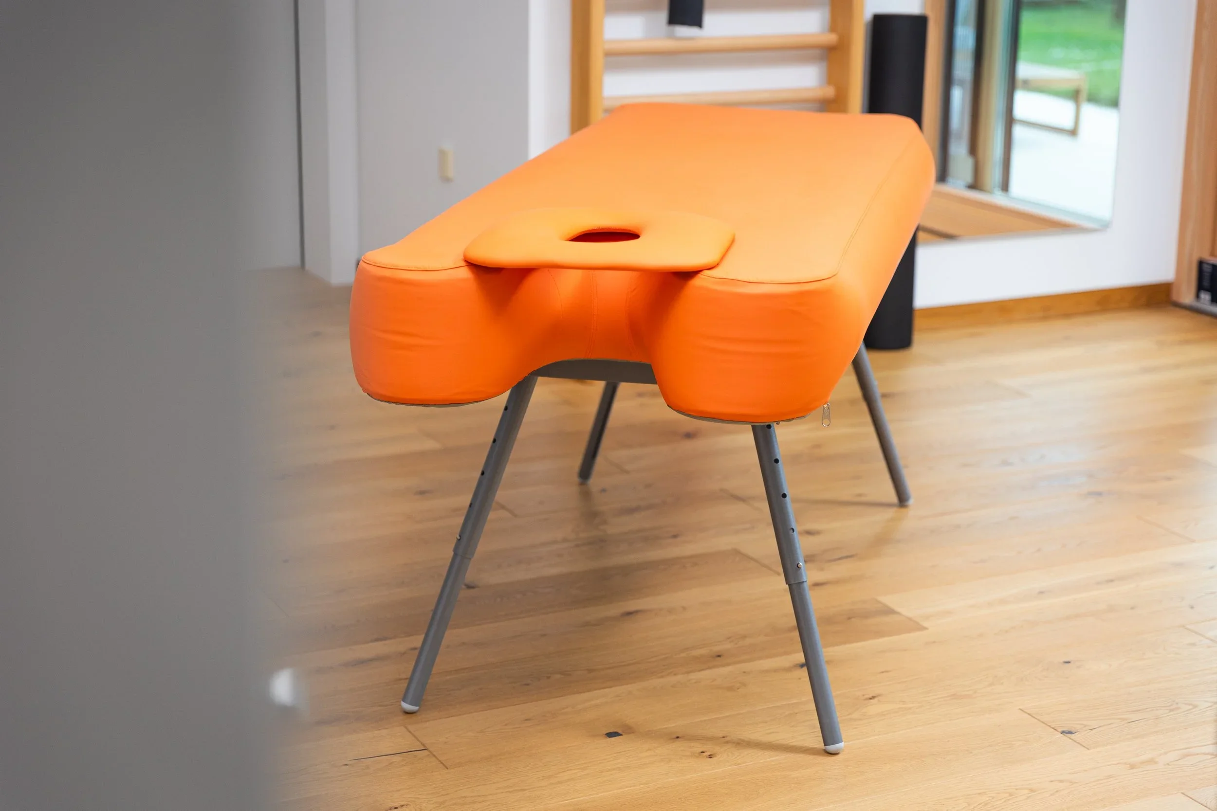 Orange massage table in a room with wooden flooring and a large window.