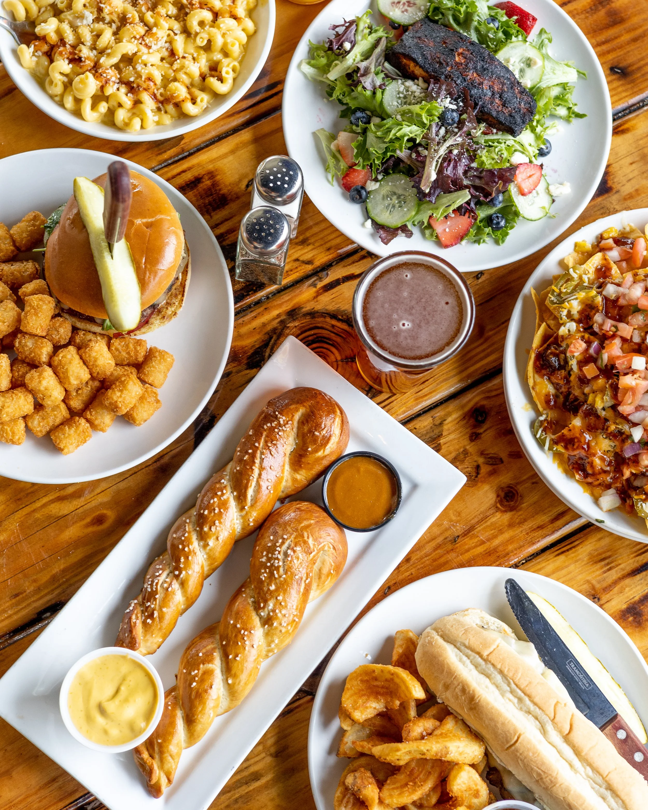 An above photo for the Vintage Brewing Company of a blackened salmon salad, chicken mac and cheese, burger and bar pretzel with a beer in the middle. 