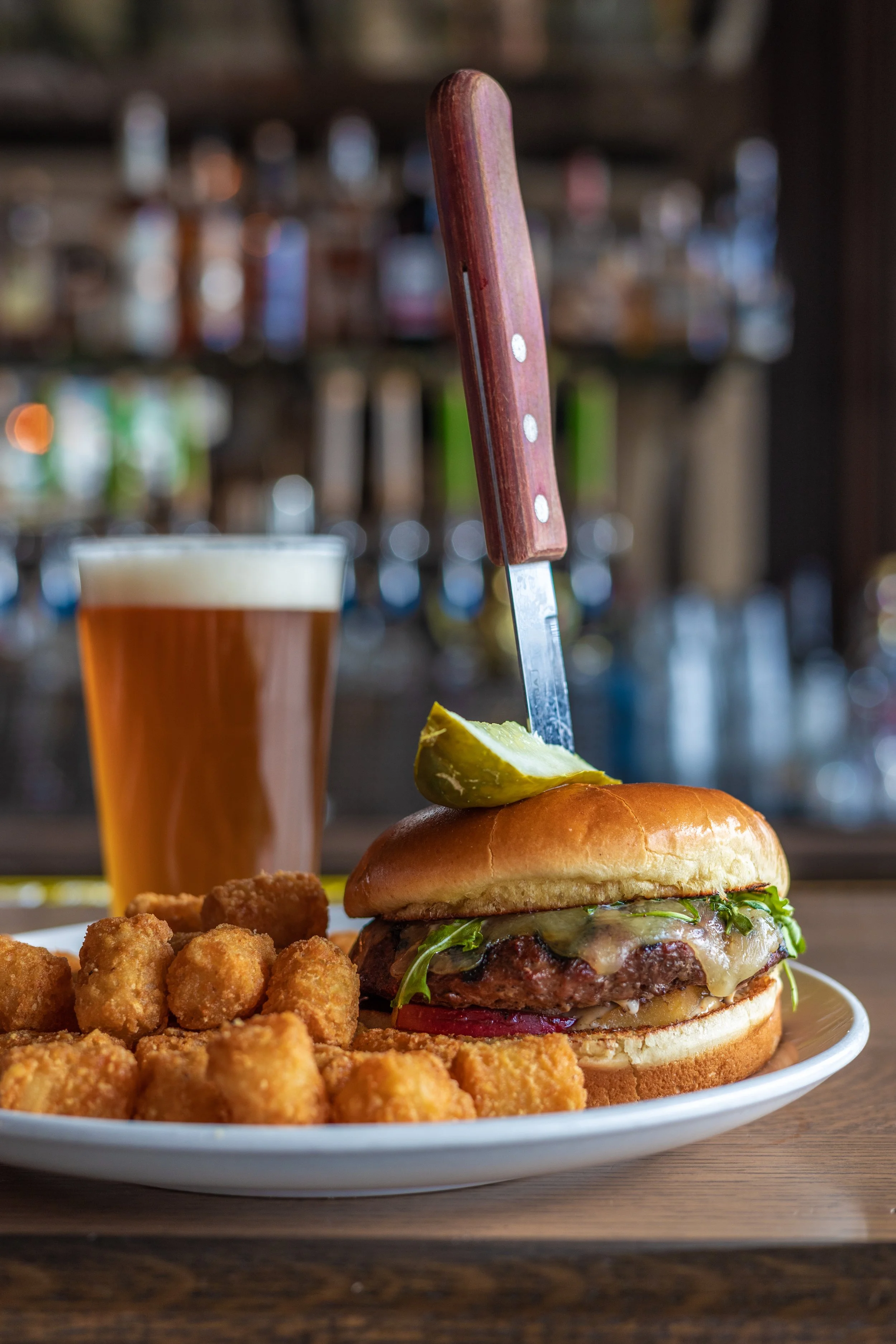 A burger with a knife in it, tater tots on the side and a beer at the vintage brewing company