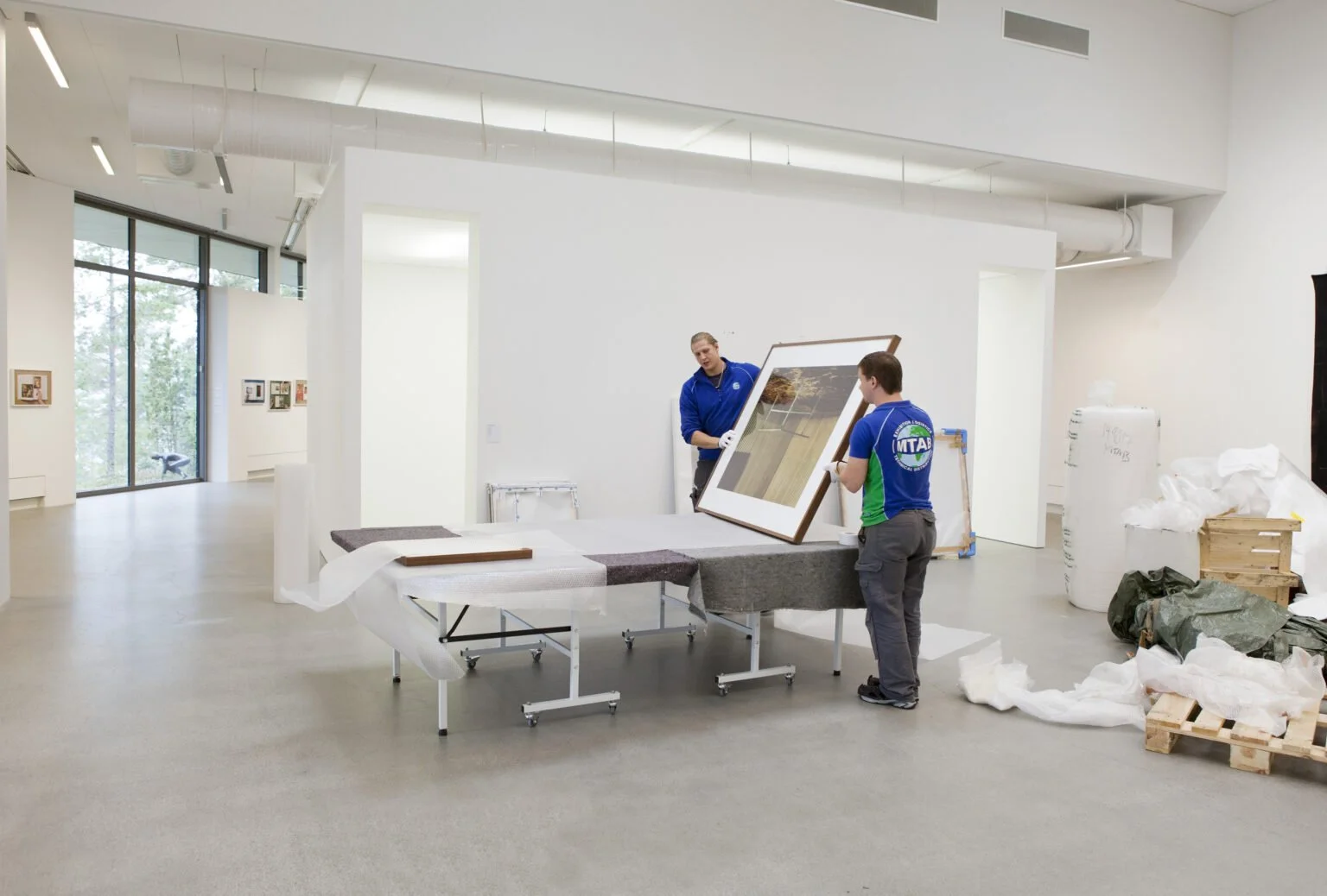 Two workers in an art gallery handling a large framed photograph or artwork on a table. The gallery has white walls and large windows. There are art pieces on the walls, and packing or construction materials on the floor.