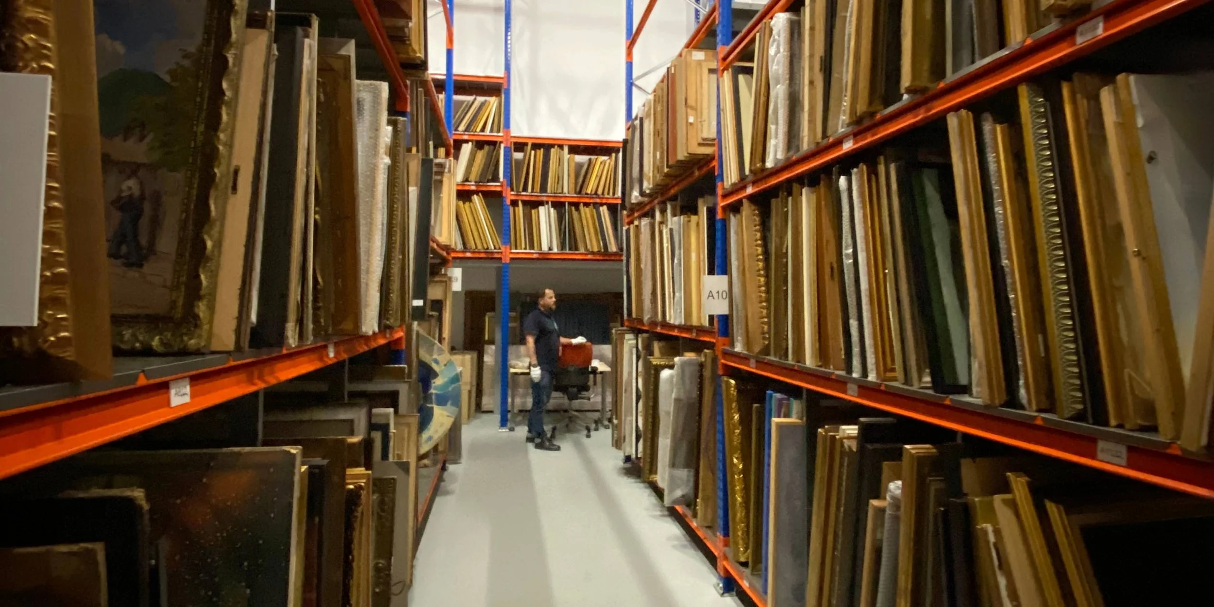 A worker in a warehouse aisle surrounded by shelves filled with framed artwork and mirrors.