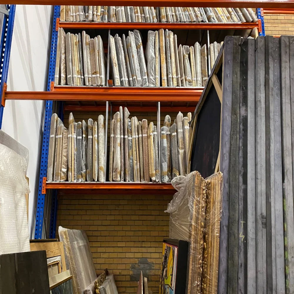 Metal shelves filled with rolled up framed artwork or posters stored vertically, some leaning against a brick wall, with additional artworks and materials stacked nearby.
