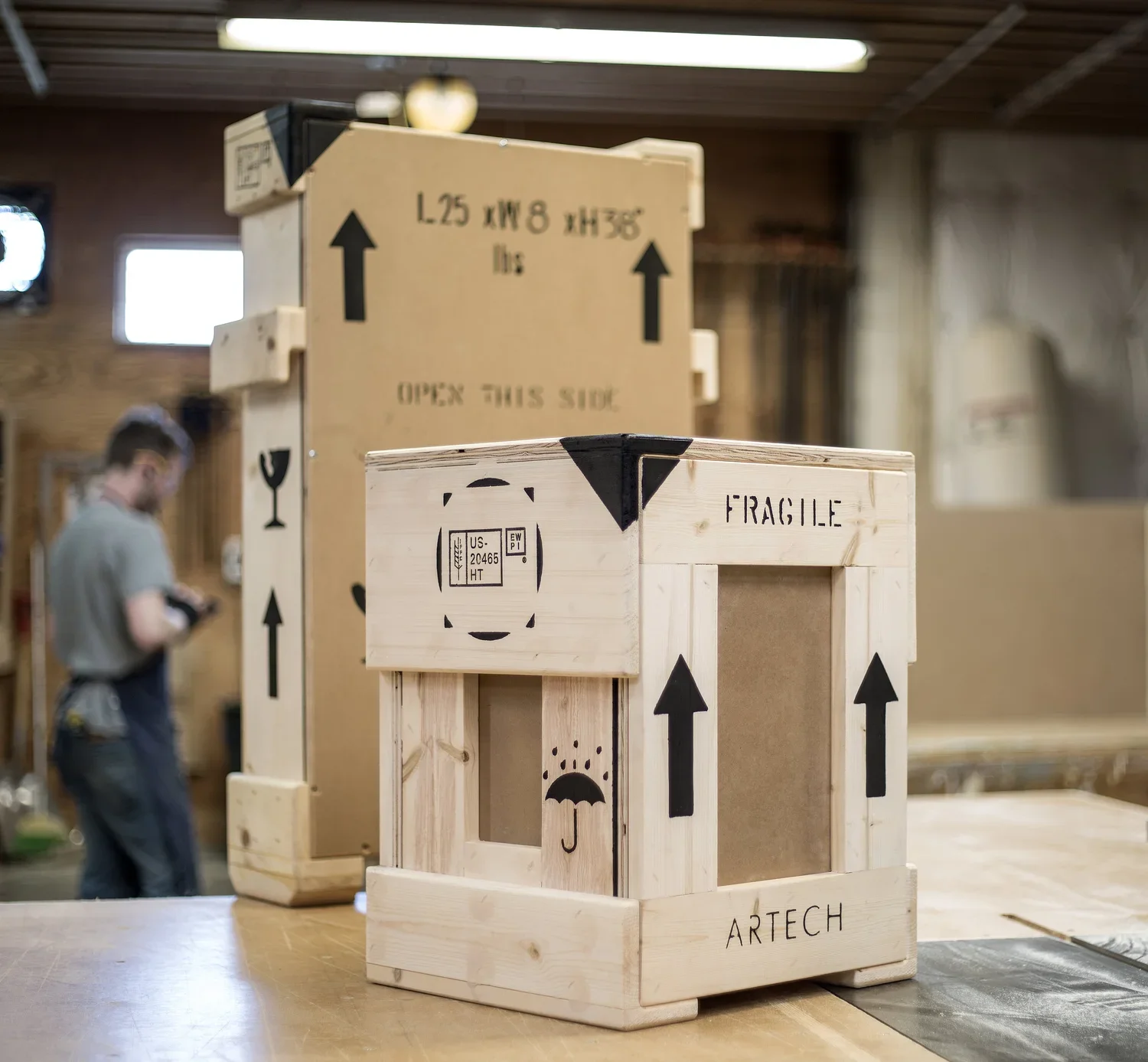Wooden crates with black arrow markings and labels such as "FRAGILE," "OPEN THIS SIDE," and "ARTECH" inside a workshop with wood walls and a person in the background.
