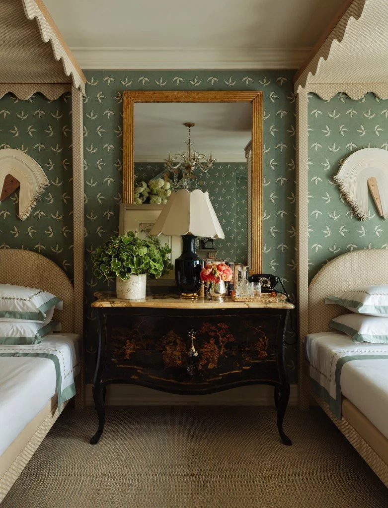 A symmetrical hotel room with two single beds, a black vintage nightstand with a floral design, topped with a lamp, plants, and decorative items, and a large mirror reflecting a chandelier and floral arrangements.