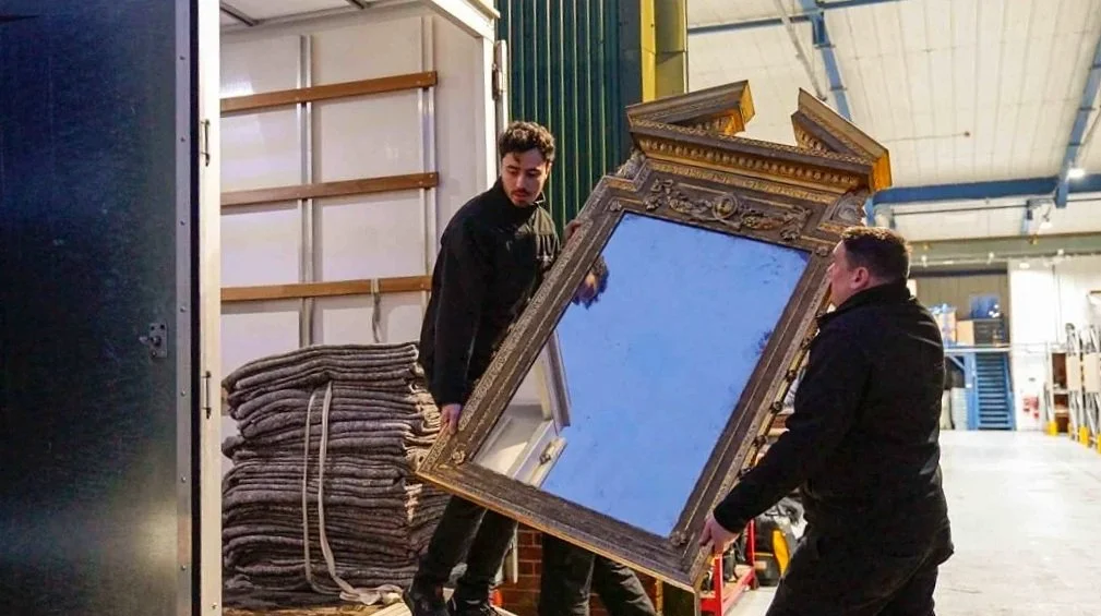 Two men are lifting a large ornate mirror with a gold frame in a warehouse.