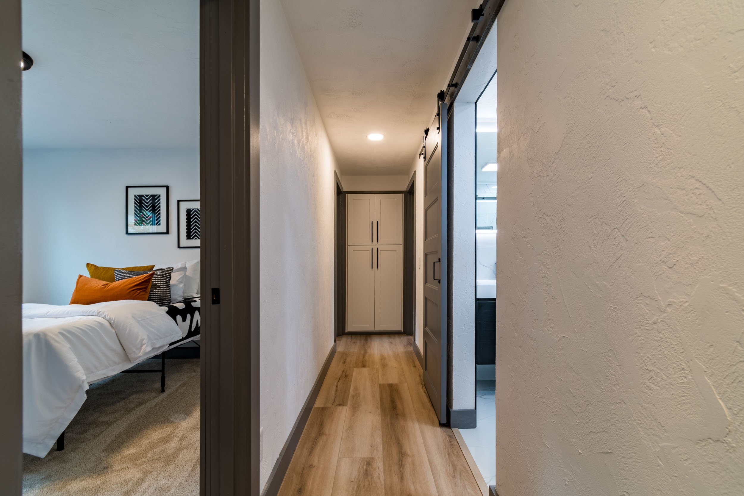 Interior hallway with wood flooring, white walls, leading to a bedroom with a bed and framed art and a bathroom with modern fixtures.