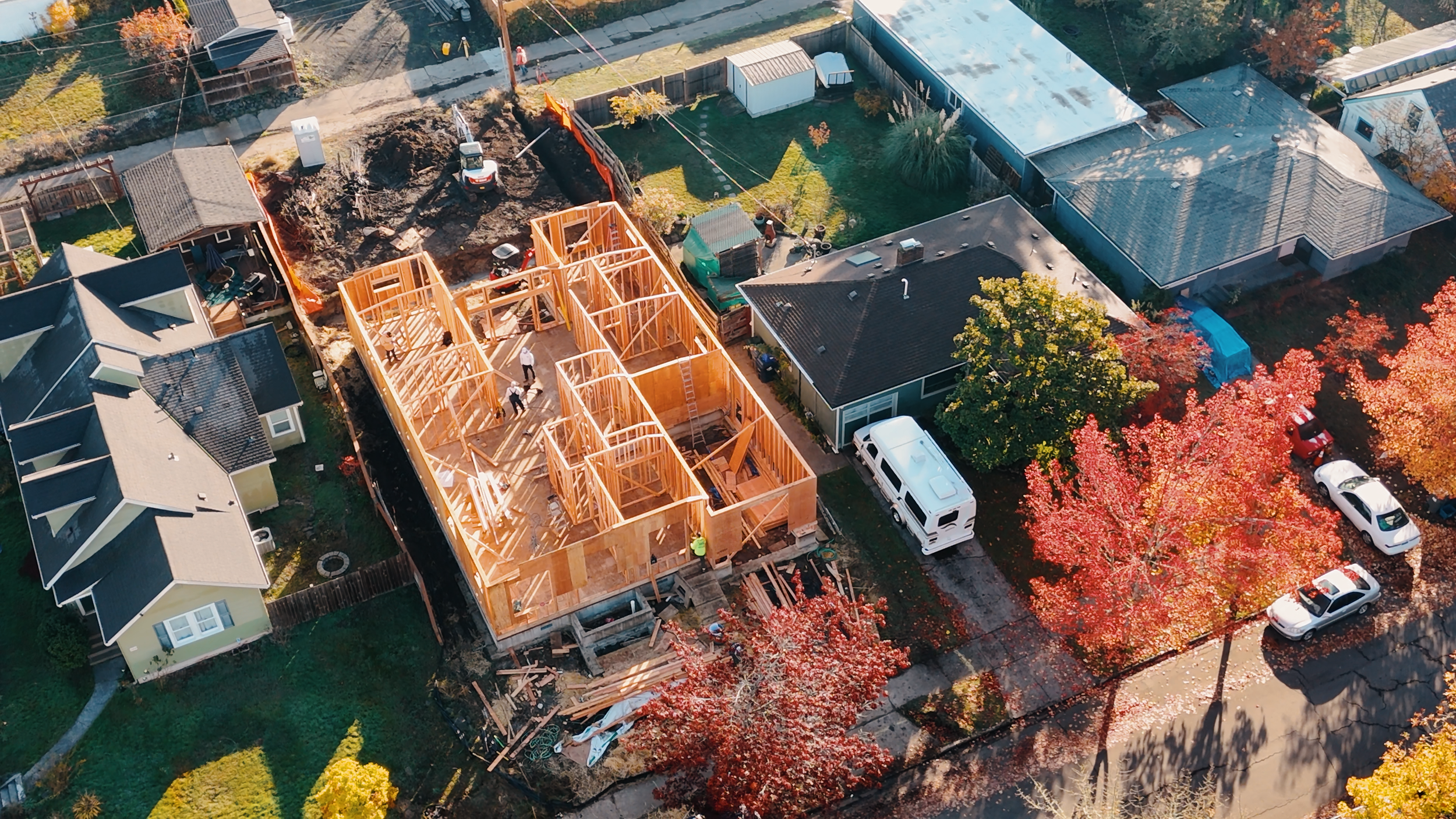 Aerial view of a residential area under construction, showing a wooden house framework, nearby homes, trees with autumn foliage, parked cars, and construction materials scattered around the site.