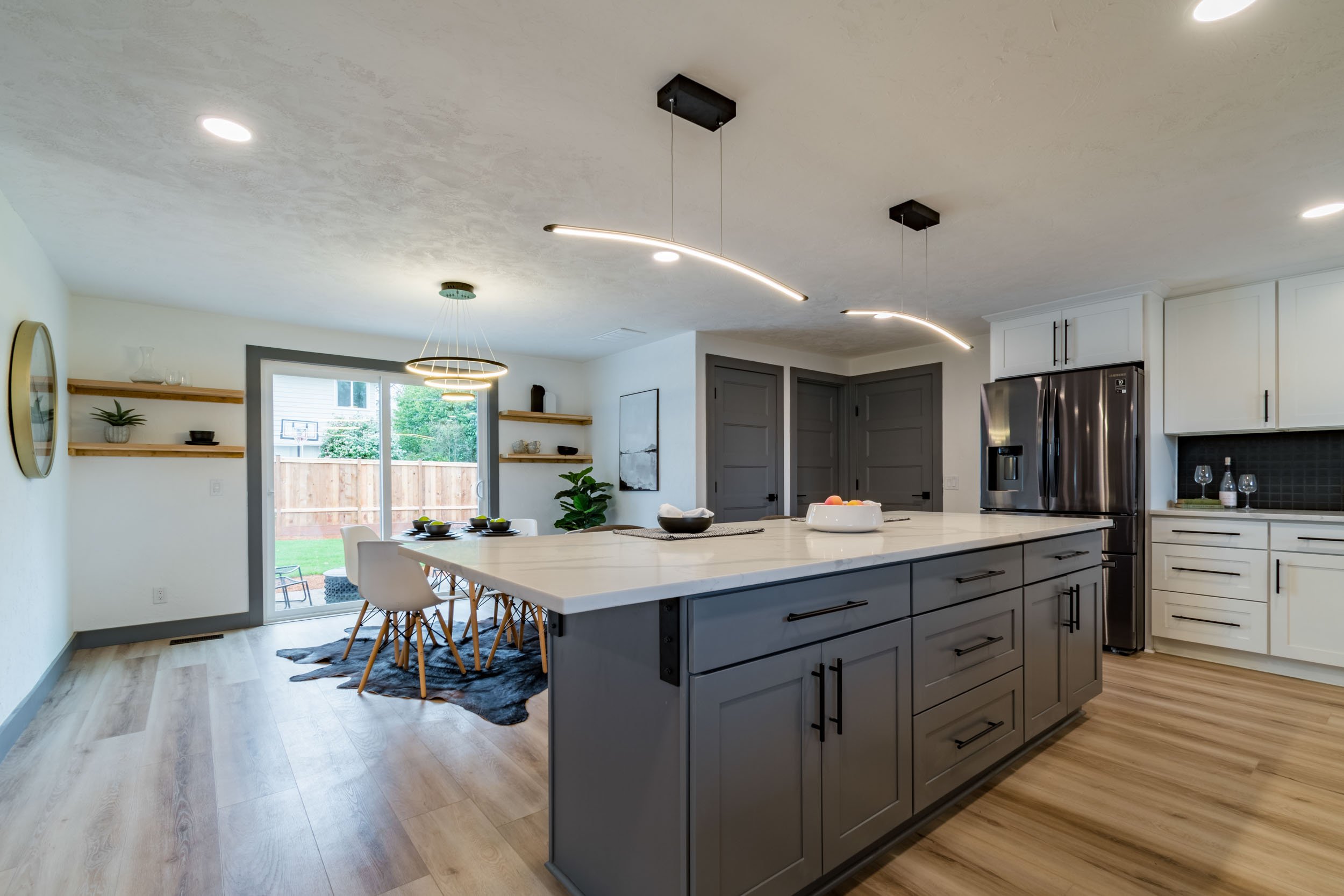 Modern kitchen and dining area with a large island, sleek pendant lighting, dark gray cabinetry, stainless steel refrigerator, and natural wood flooring. The dining space includes a table with white chairs and sliding glass doors leading to a patio.