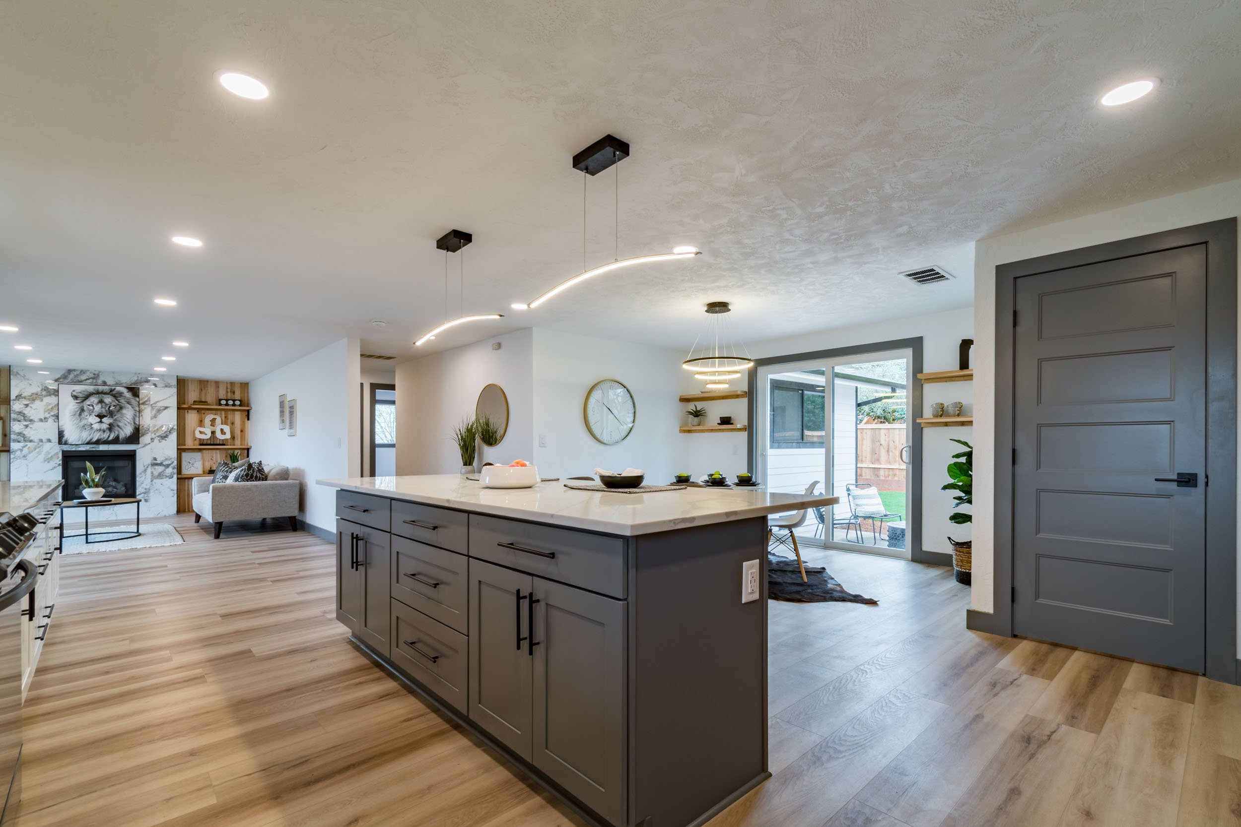 Modern open-concept kitchen and living area with gray cabinetry, wood flooring, and stylish lighting. Features large kitchen island, sliding glass doors to patio, and cozy sitting area with fireplace.