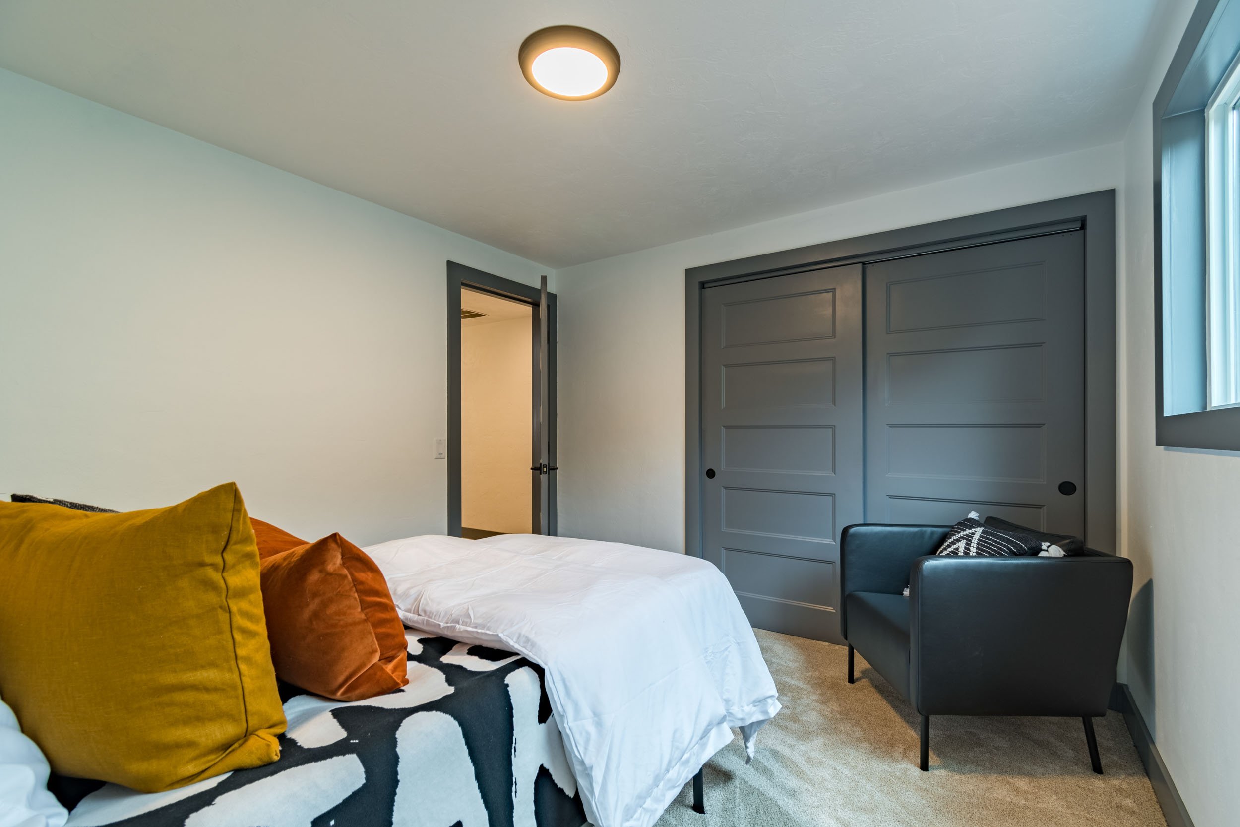 Modern bedroom with gray walls and closet doors, featuring a bed with yellow and orange pillows, a black armchair, and a window with natural light.
