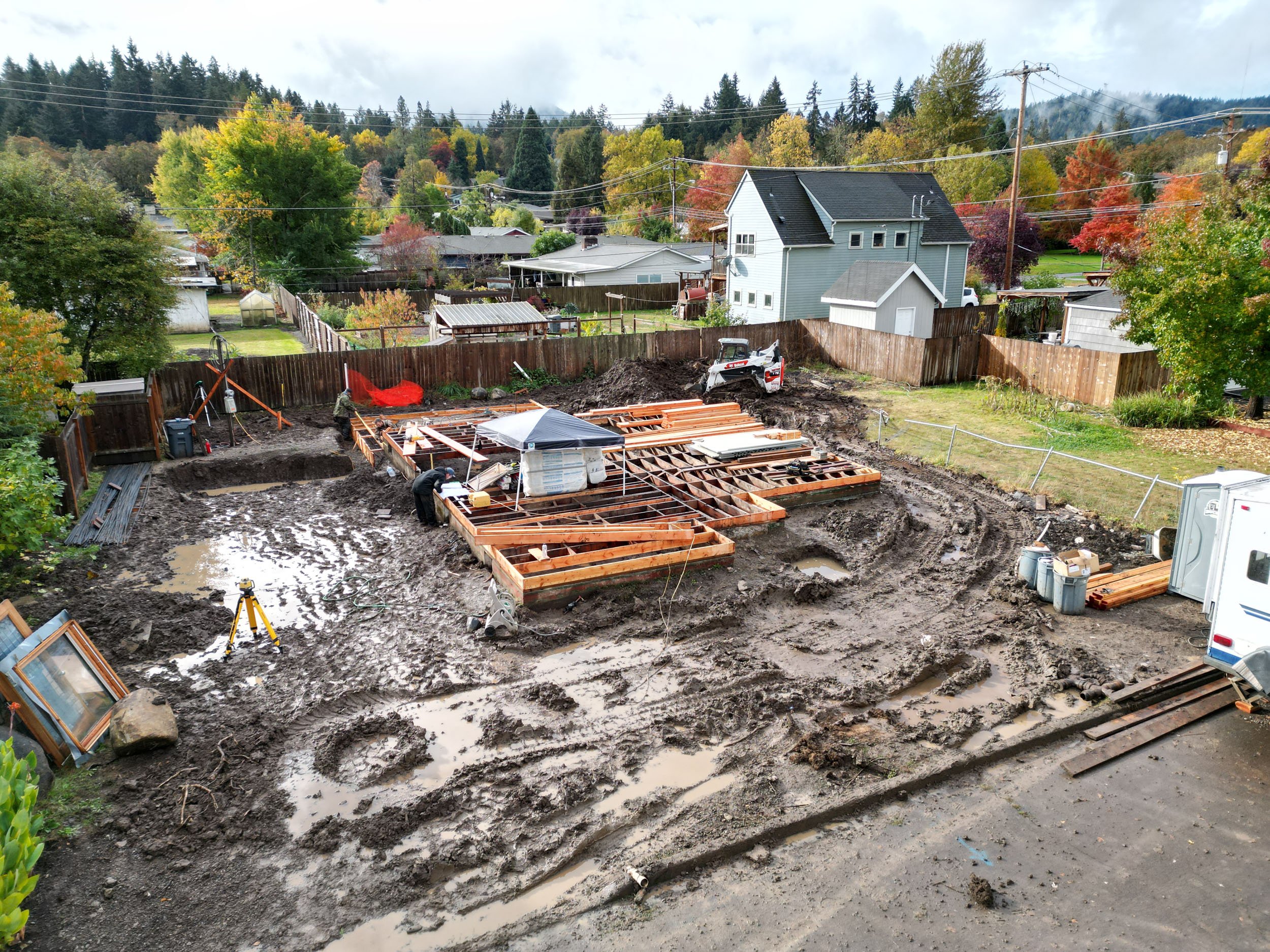 Construction site with muddy ground and wooden foundation frames in a residential area, surrounded by trees and houses.
