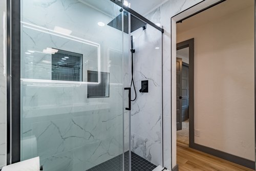 Modern bathroom with a glass shower enclosure, marble tiles, and a rainfall showerhead, next to a wooden floor hallway.