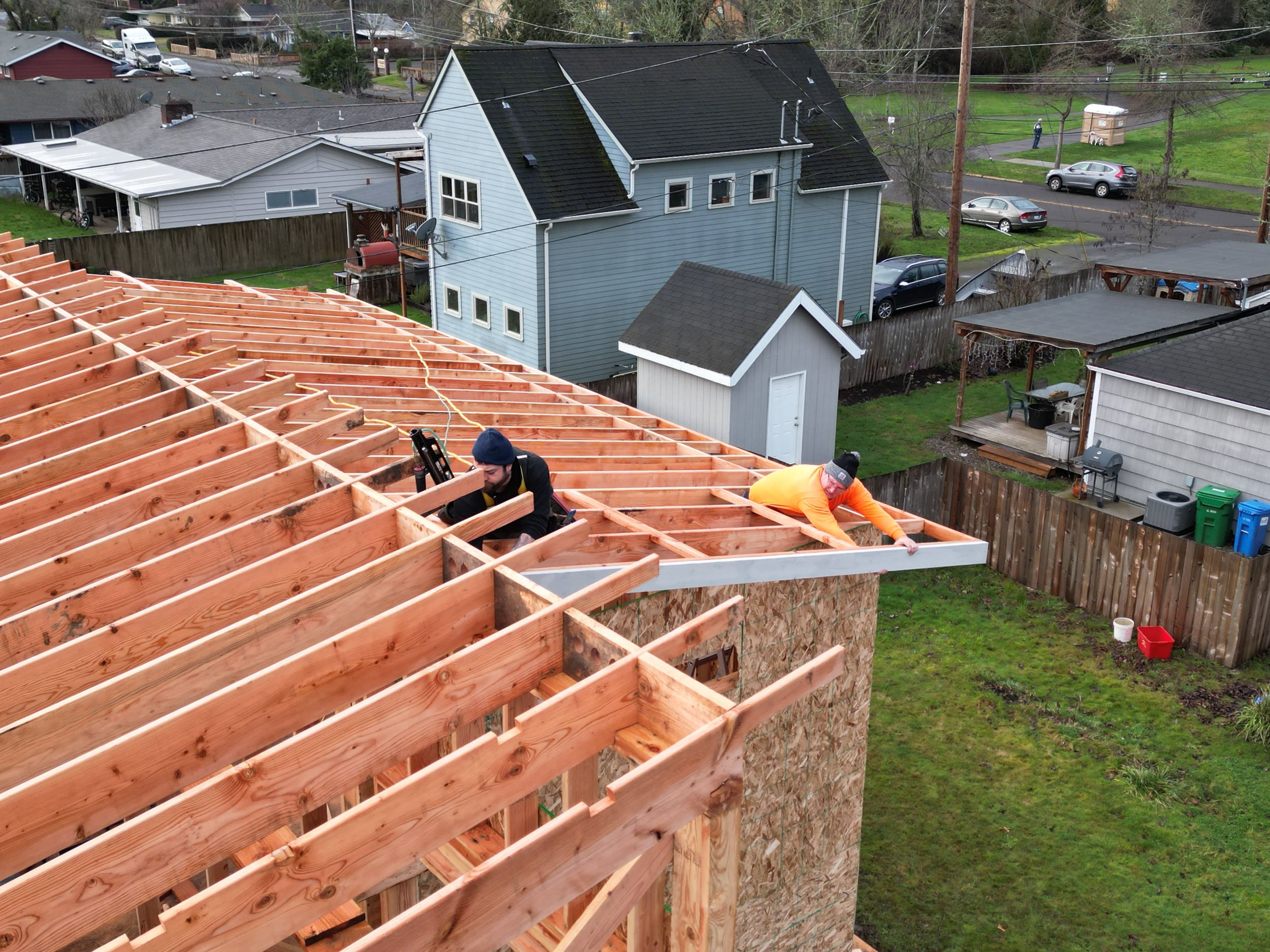 Two construction workers on a wooden roof frame of a building under construction, surrounded by residential houses and yards.