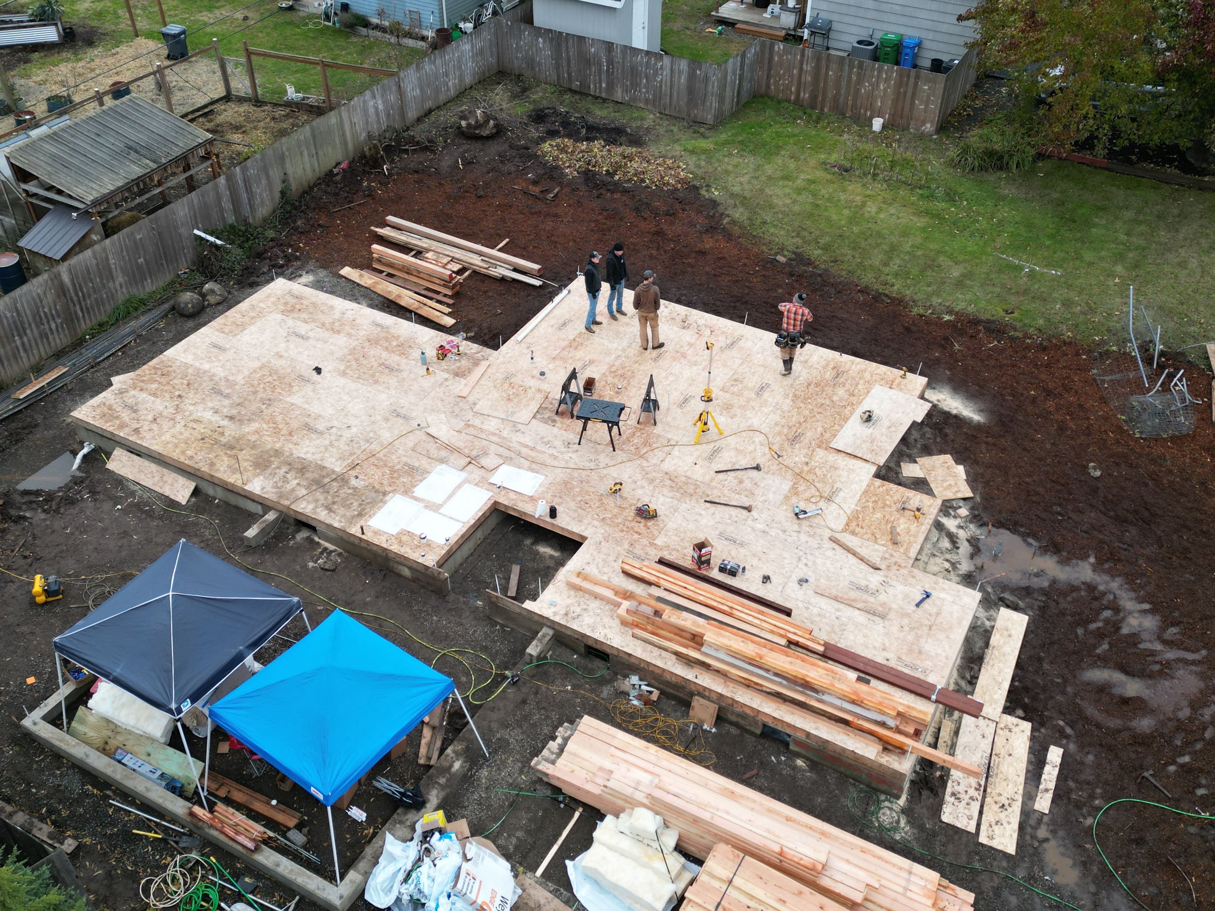 Aerial view of a construction site with a wooden foundation being built in a backyard. Several people are working on the structure, surrounded by tools, wooden planks, and two small tents. The area is fenced and the ground is partially covered with g