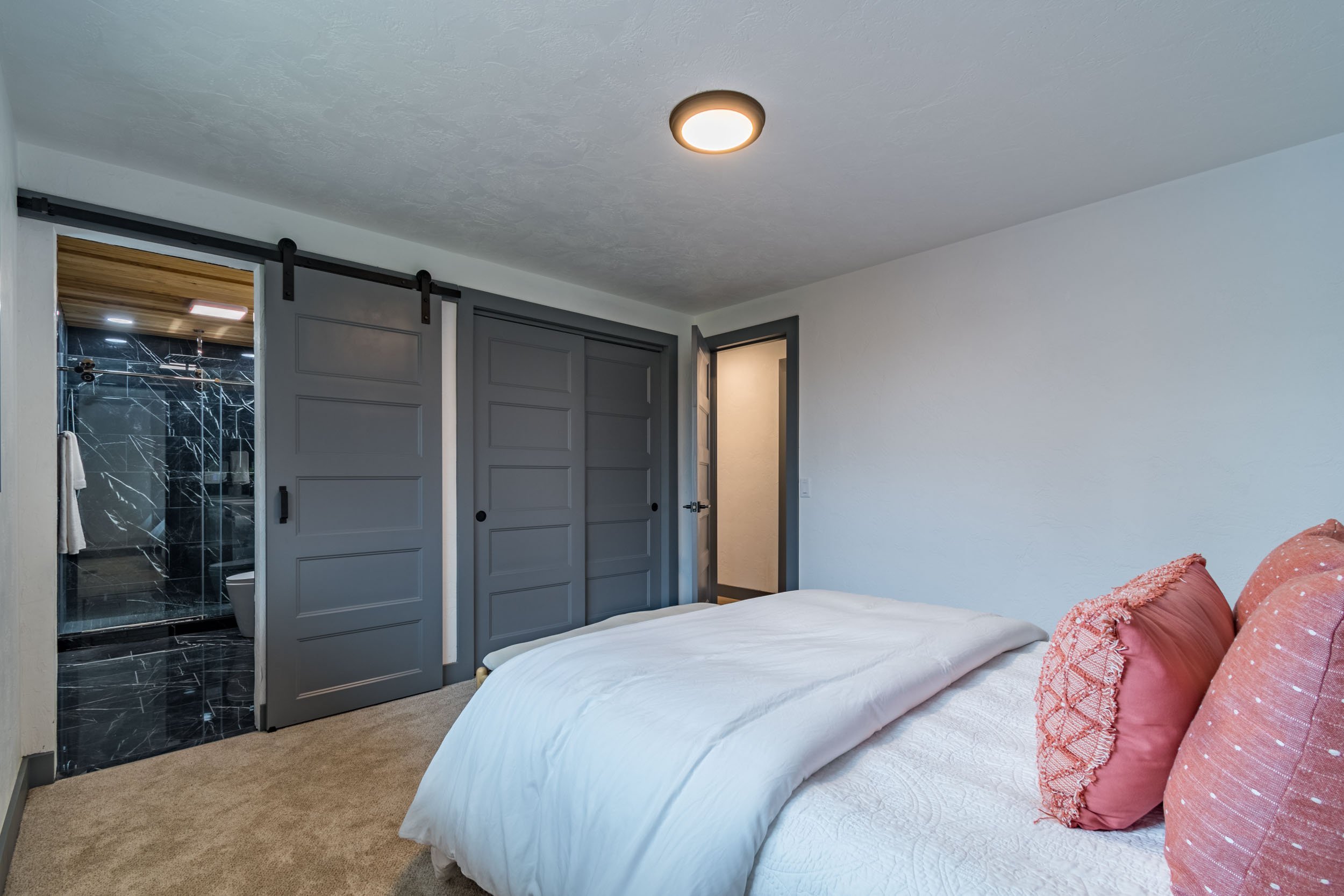 Modern bedroom with a bed, white comforter, and pink pillows, featuring gray sliding barn doors leading to a black marble bathroom.