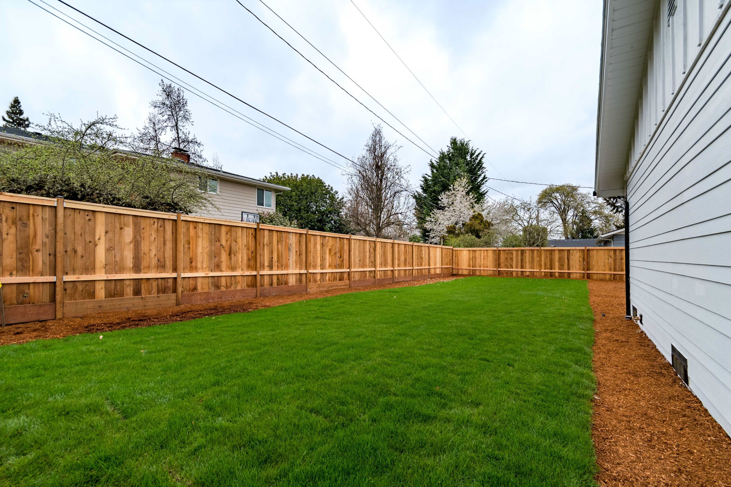 Backyard with wooden fence, green grass lawn, and house siding on the right.