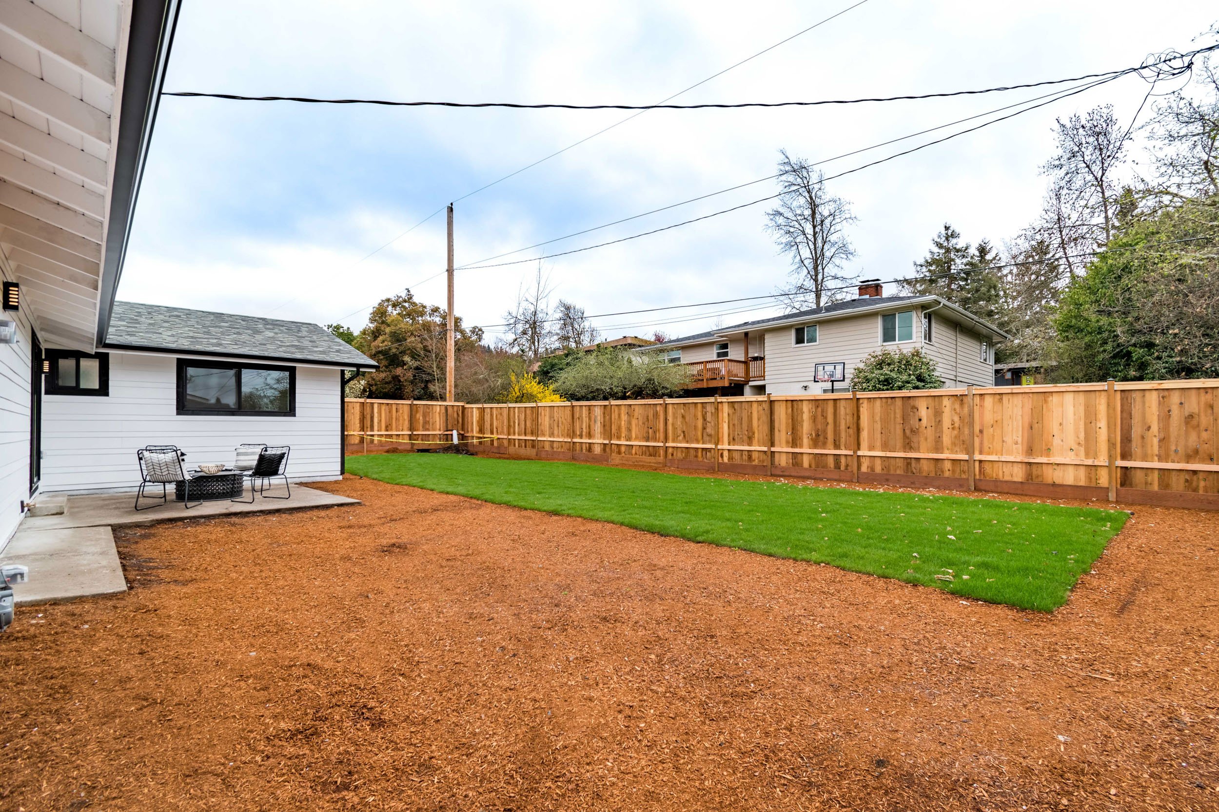 Backyard with wooden fence, grassy area, gravel path, and patio with chairs next to a white house.