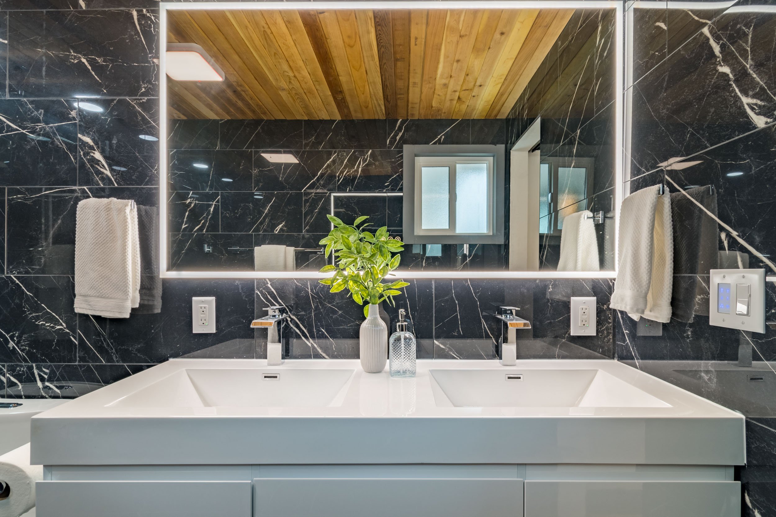 Modern bathroom with a double sink vanity, illuminated mirror, black marble walls, wooden ceiling, and a decorative plant in a vase.