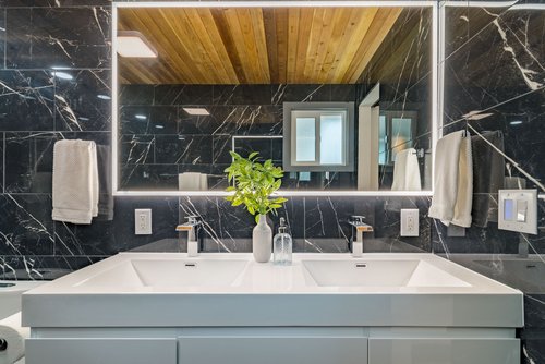 Modern bathroom with a double sink vanity, illuminated mirror, black marble walls, wooden ceiling, and a decorative plant in a vase.