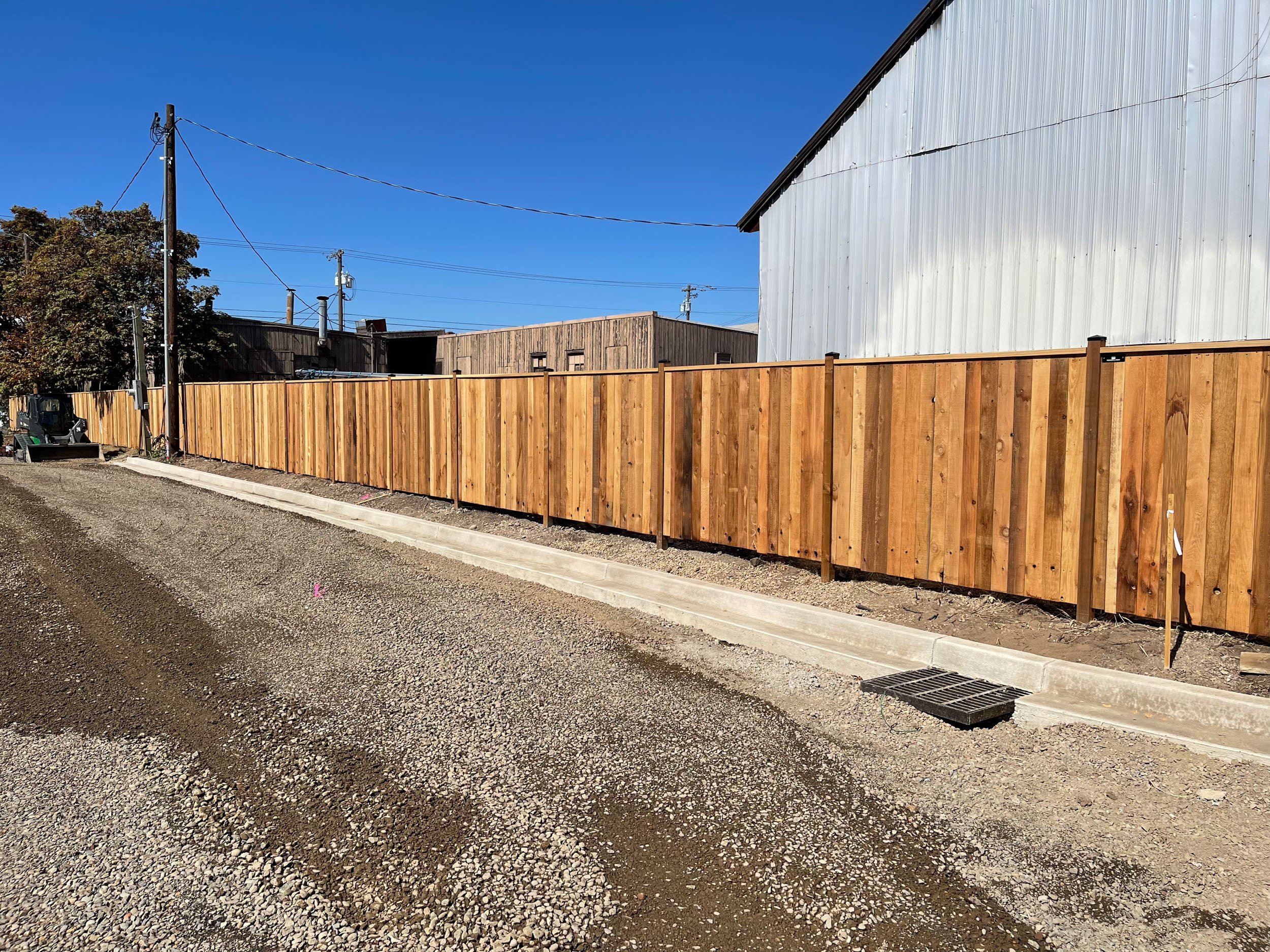 Long wooden fence next to an industrial building and gravel road under clear blue sky.