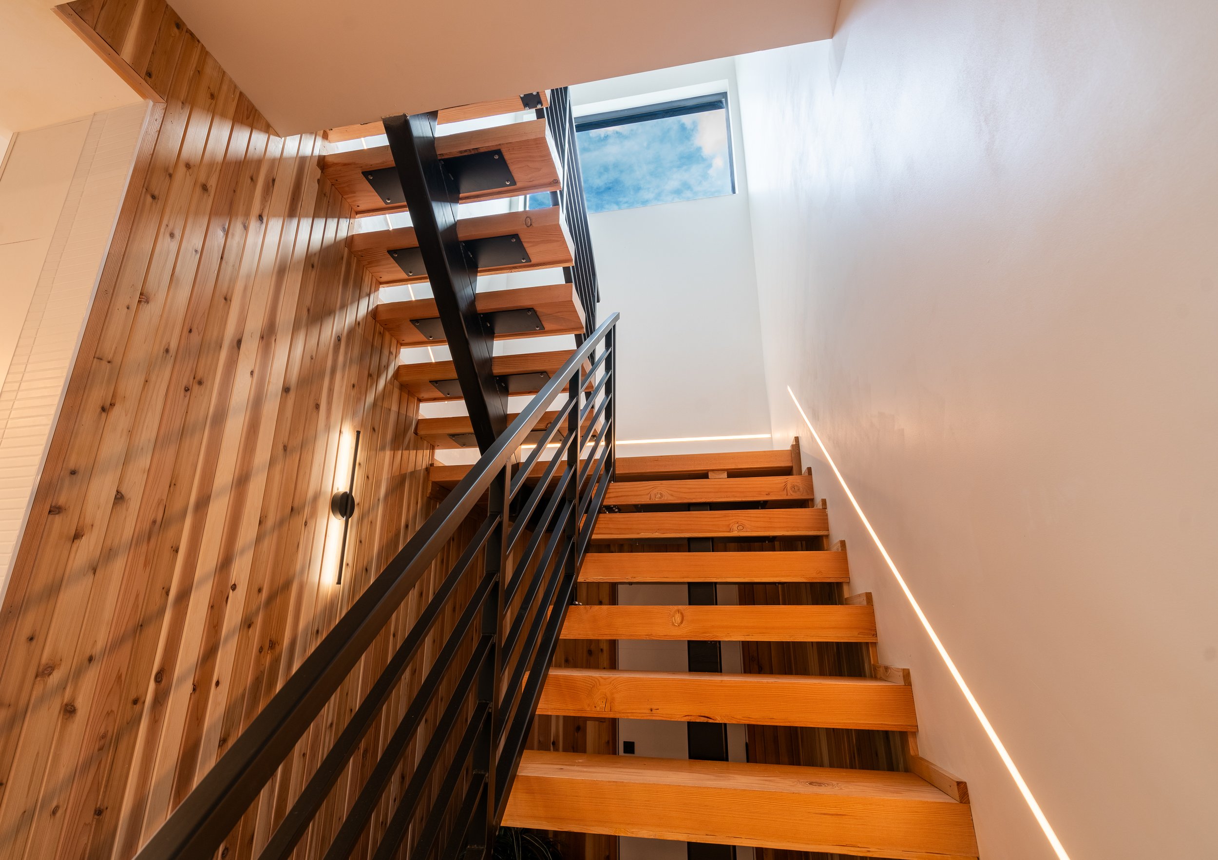 Modern wooden staircase with metal railings, featuring a wooden wall and a skylight showing blue sky.