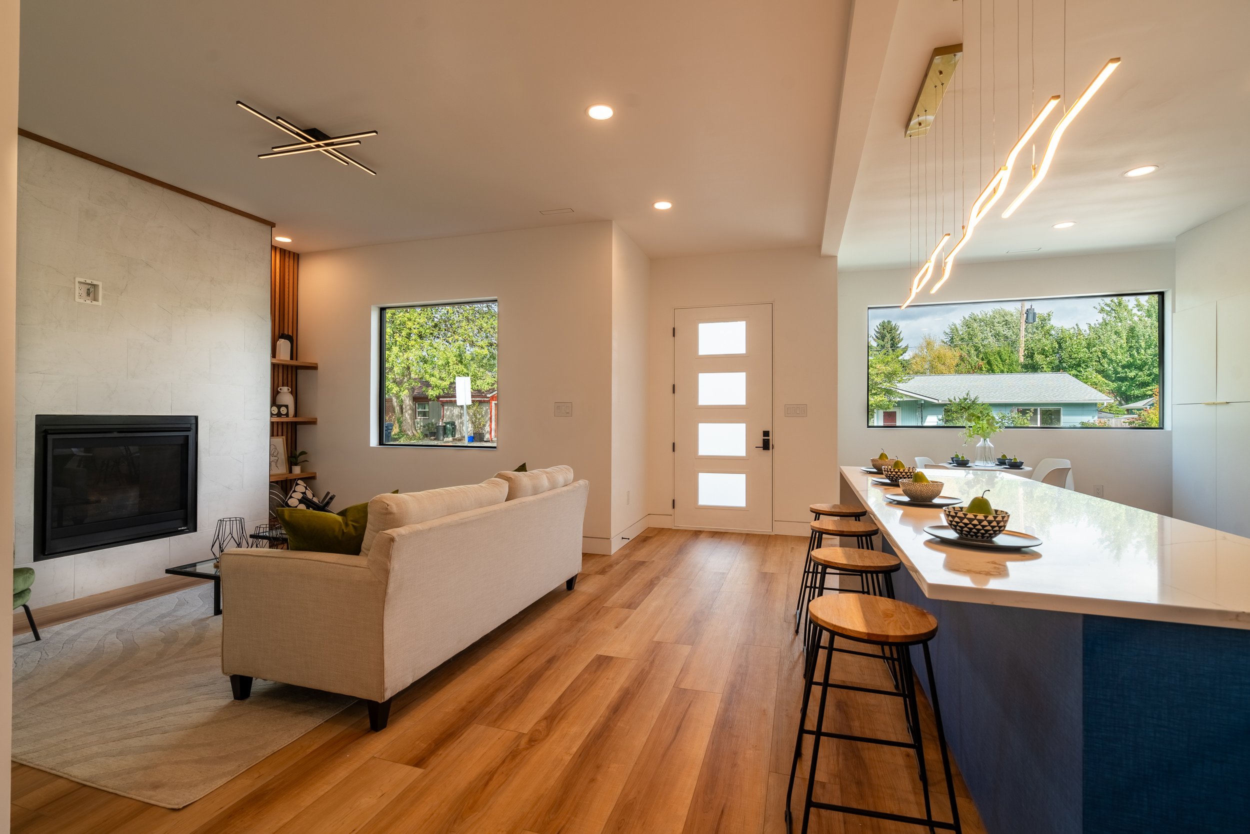 Modern open concept living room and kitchen with hardwood floors, light beige couch, wall-mounted fireplace, large windows with greenery outside, white door with frosted glass panels, and a kitchen island with bar stools and pendant lights above.