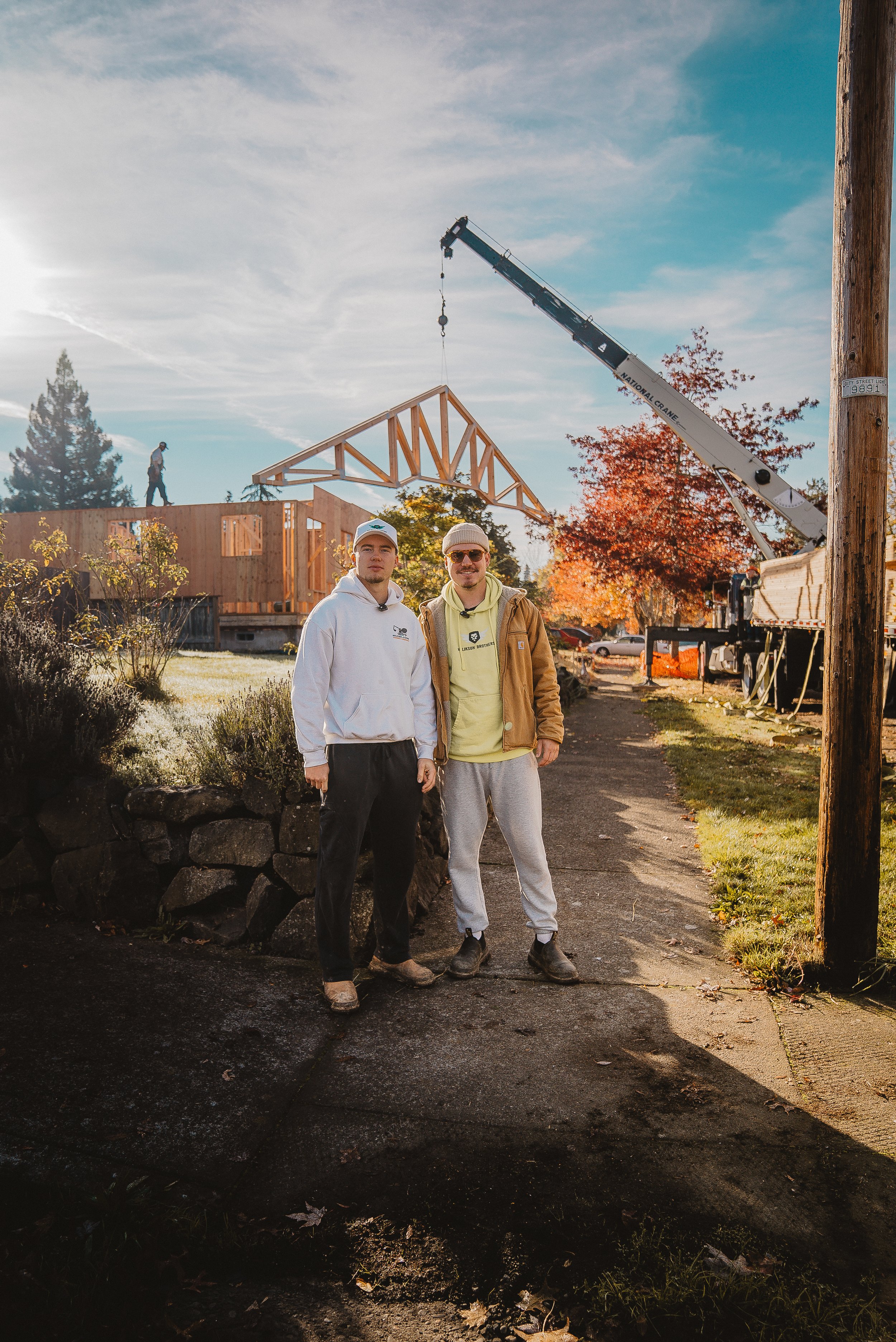 Two men posing in front of a house construction site with a crane lifting a roof truss and workers on the structure.