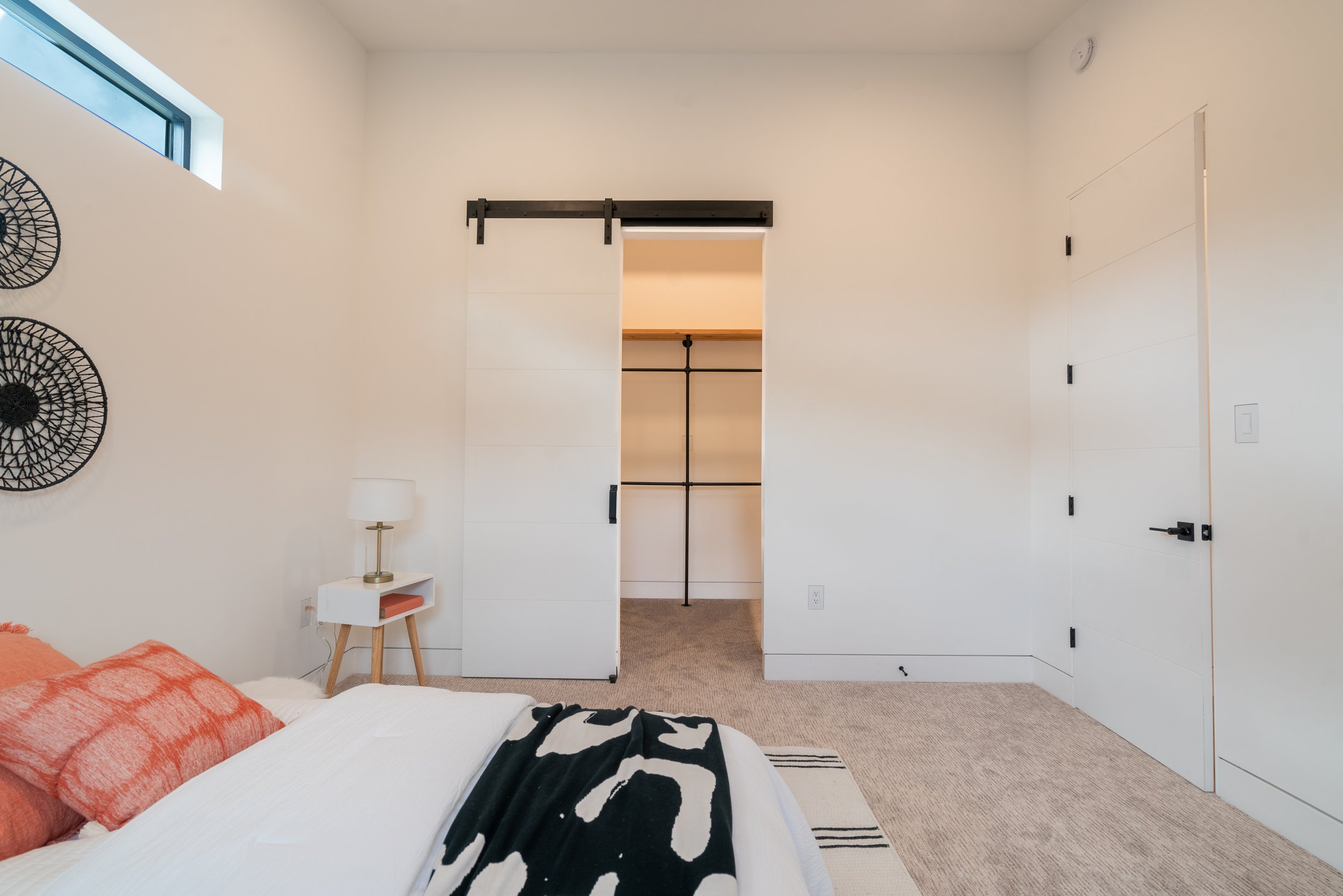 Minimalist bedroom with white walls, a bed with orange and black accents, a small wooden nightstand with a lamp, black decorative wall art, and a sliding barn door leading to a closet.