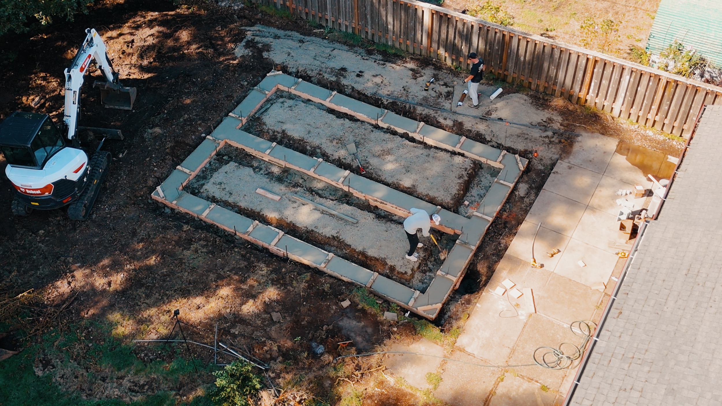 Aerial view of a backyard construction site showing concrete foundation forms, two workers, an excavator, and a wooden fence.