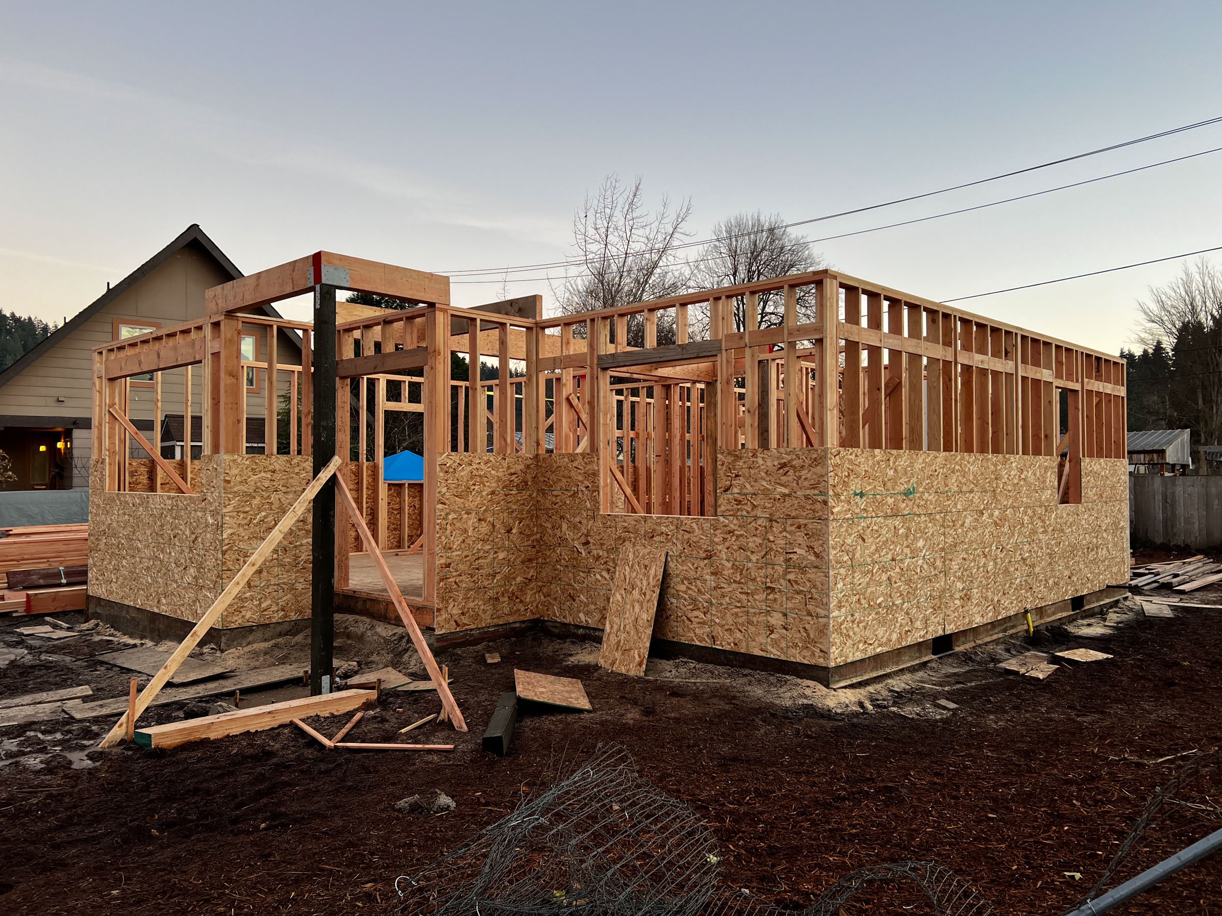 Wooden structure under construction of a house, with framing visible, surrounded by a dirt yard with construction materials scattered around.