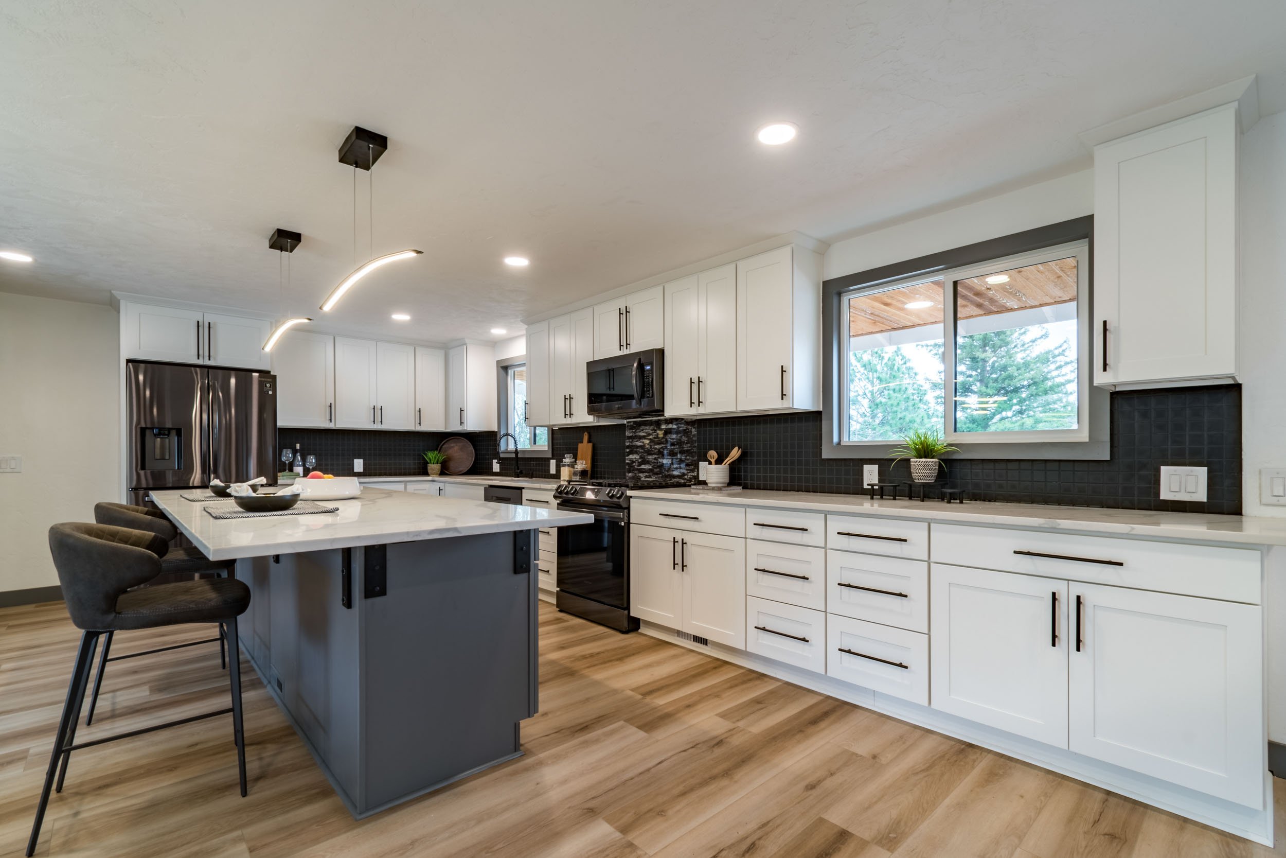 Modern kitchen with white cabinets, black backsplash, stainless steel appliances, and a large island with bar stools. Wooden flooring and contemporary pendant lights complete the design.
