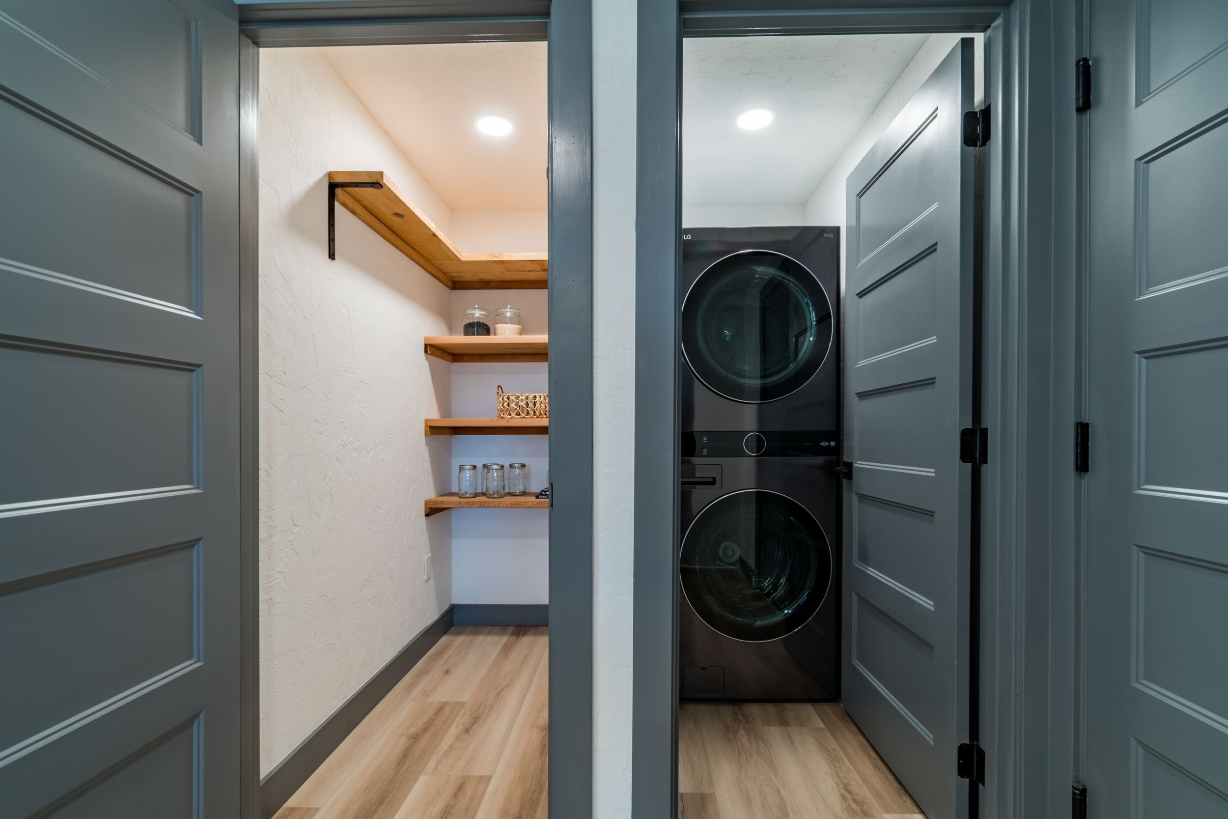 Interior of a small utility room with a stackable washer and dryer on the right, and wooden shelves with jars and containers on the left.