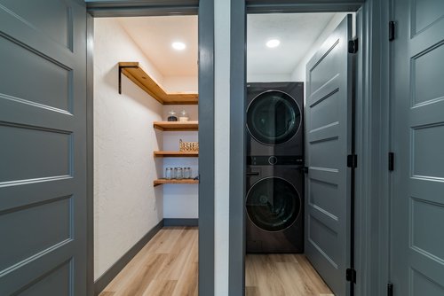 Interior of a small utility room with a stackable washer and dryer on the right, and wooden shelves with jars and containers on the left.