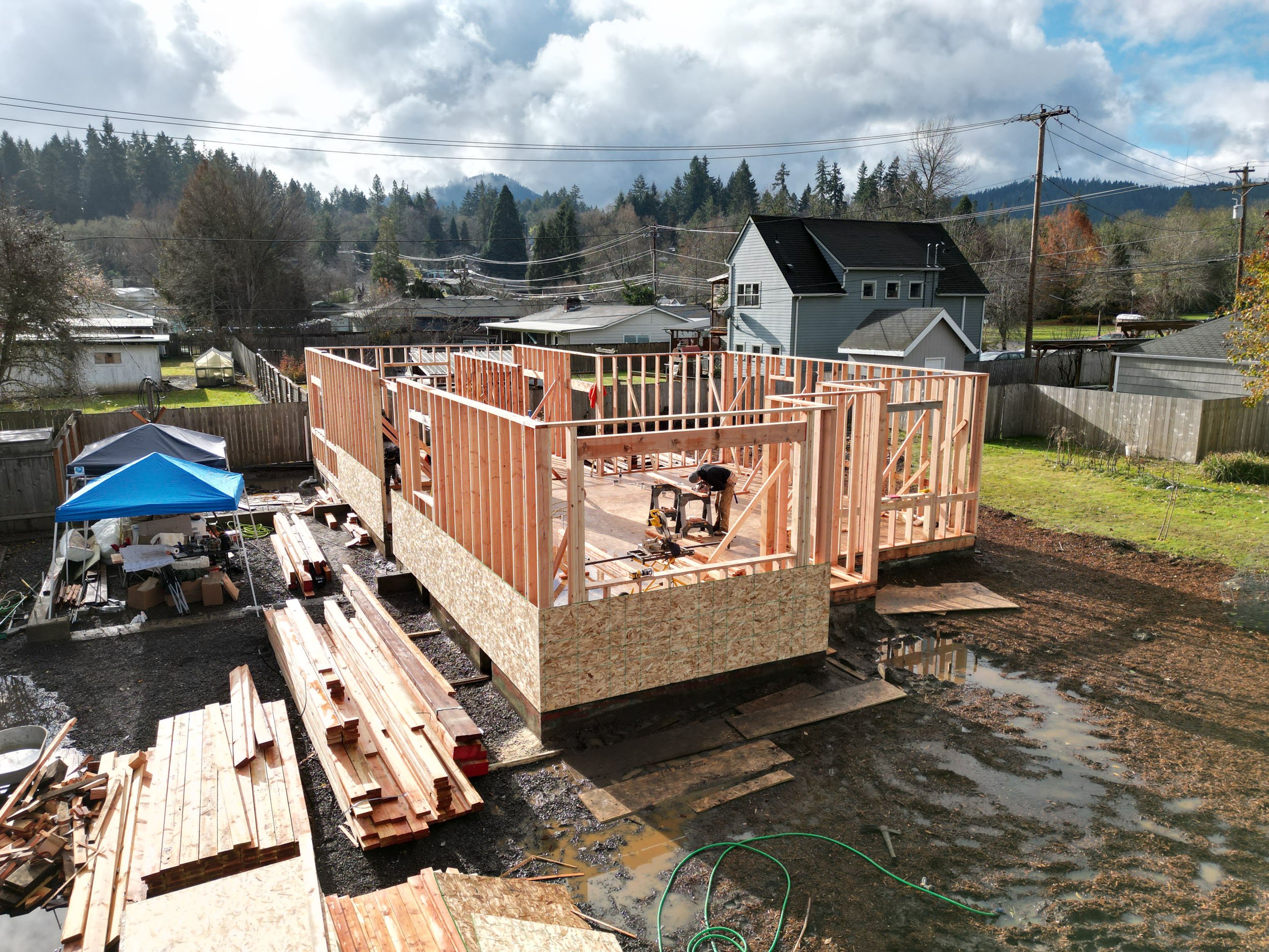 Building construction site with partially framed house, wooden beams, and a blue canopy tent nearby.