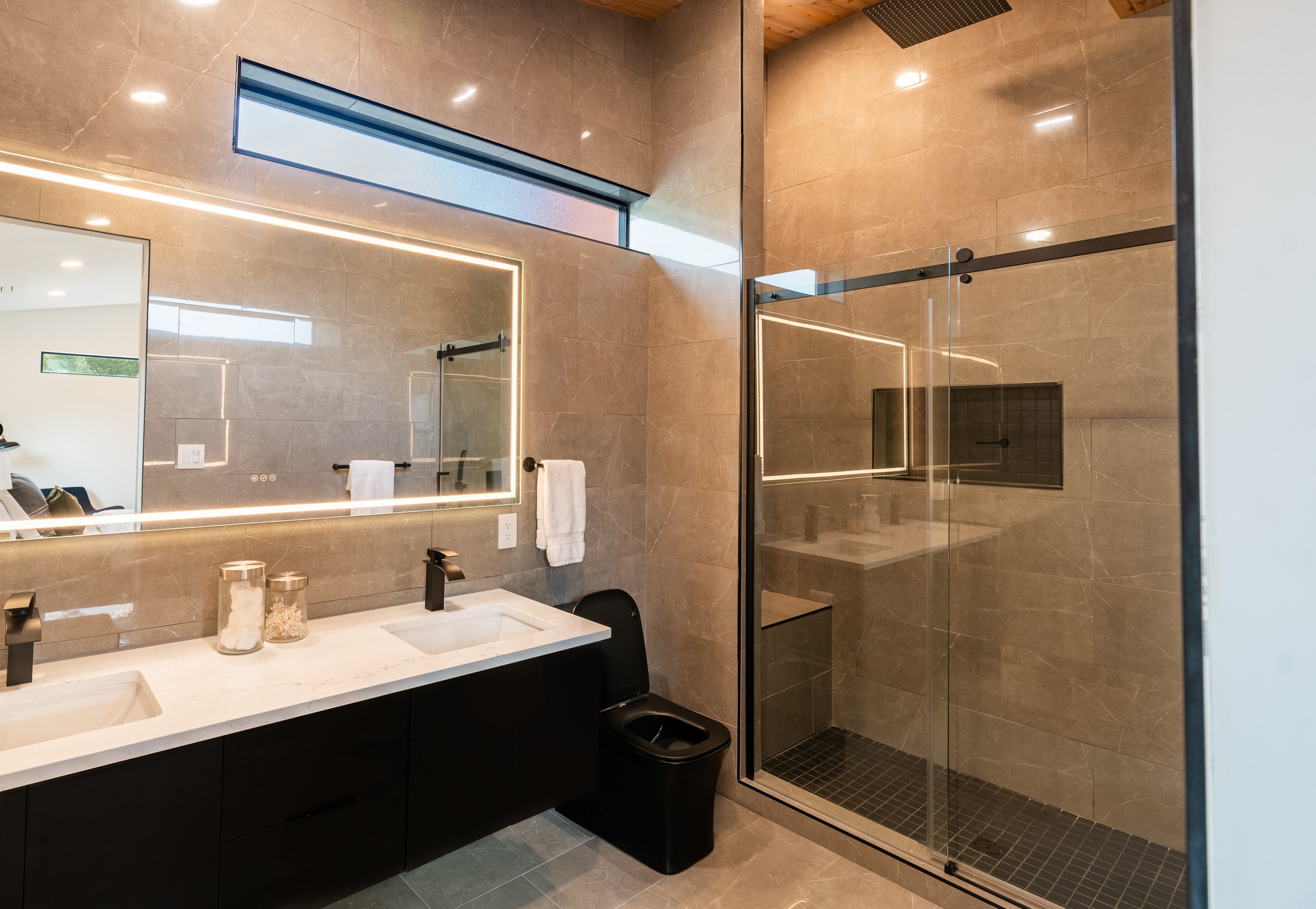 Modern bathroom with double vanity, illuminated mirror, glass-enclosed shower, and black toilet.