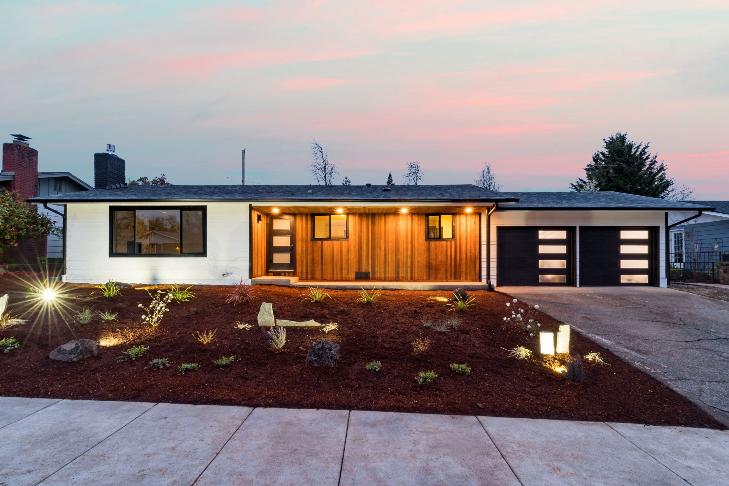 Modern single-story home with wooden siding, large windows, and a double garage at dusk, surrounded by landscaped yard and soft lighting.
